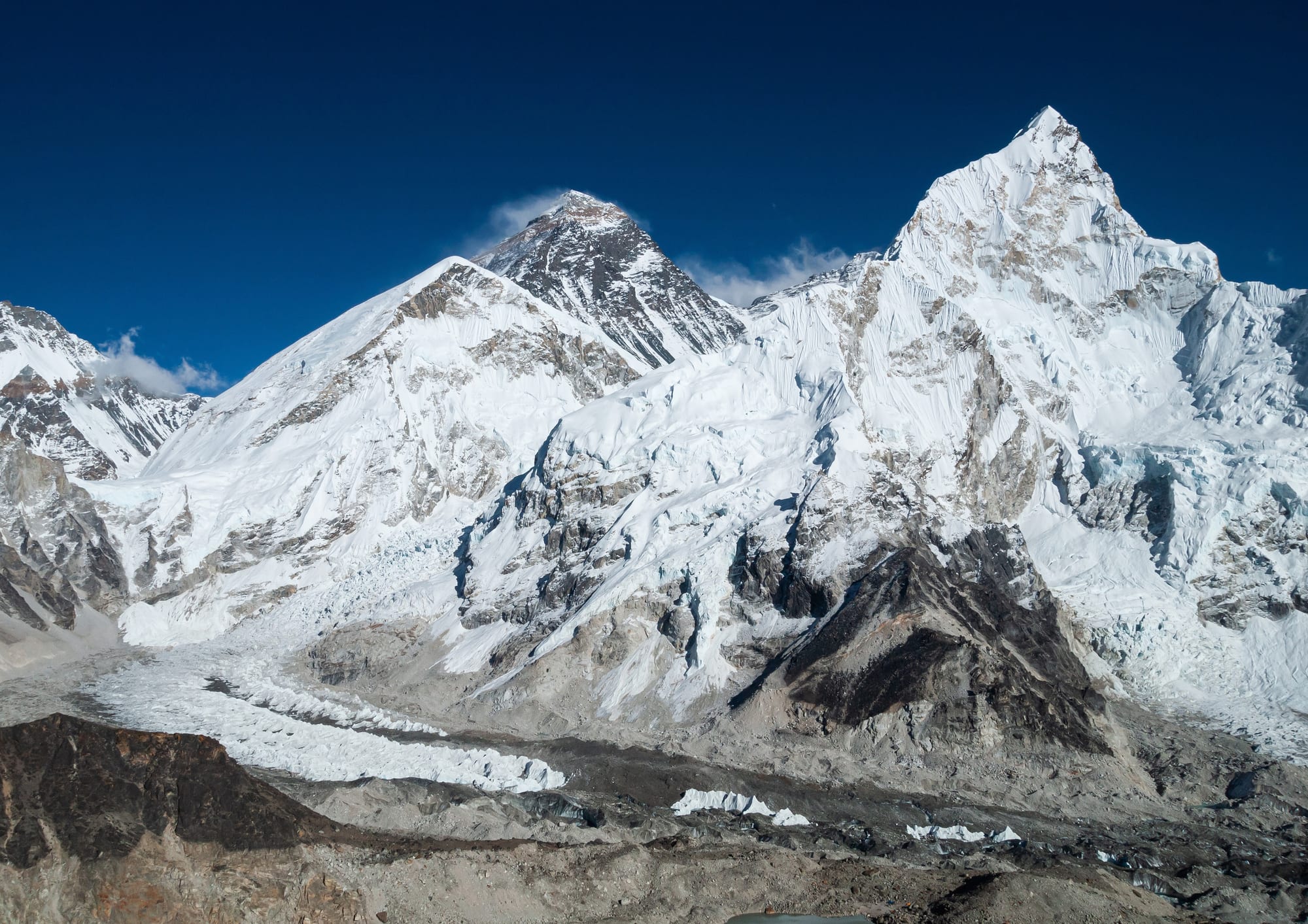 A dark rocky summit pyramid rising behind a great snow-covered ridge, with a glacier and icefall flowing out below under a deep blue sky