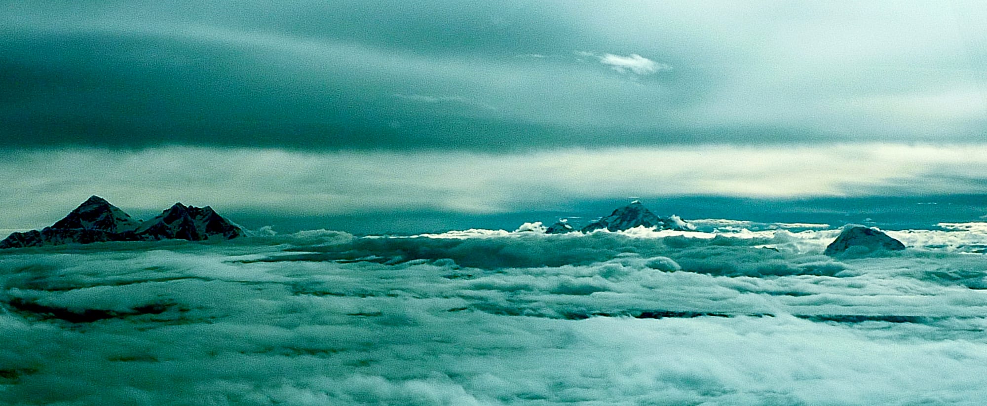 Several dark, pointed mountain summits rising above a sea of white cloud under a grey-green sky