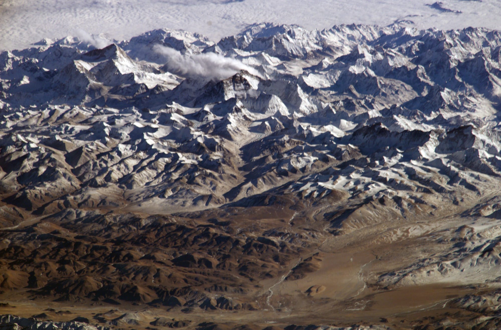 A vast field of snow-streaked, jagged peaks seen from far above, with brown, drier ground in the foreground and cloud on the horizon