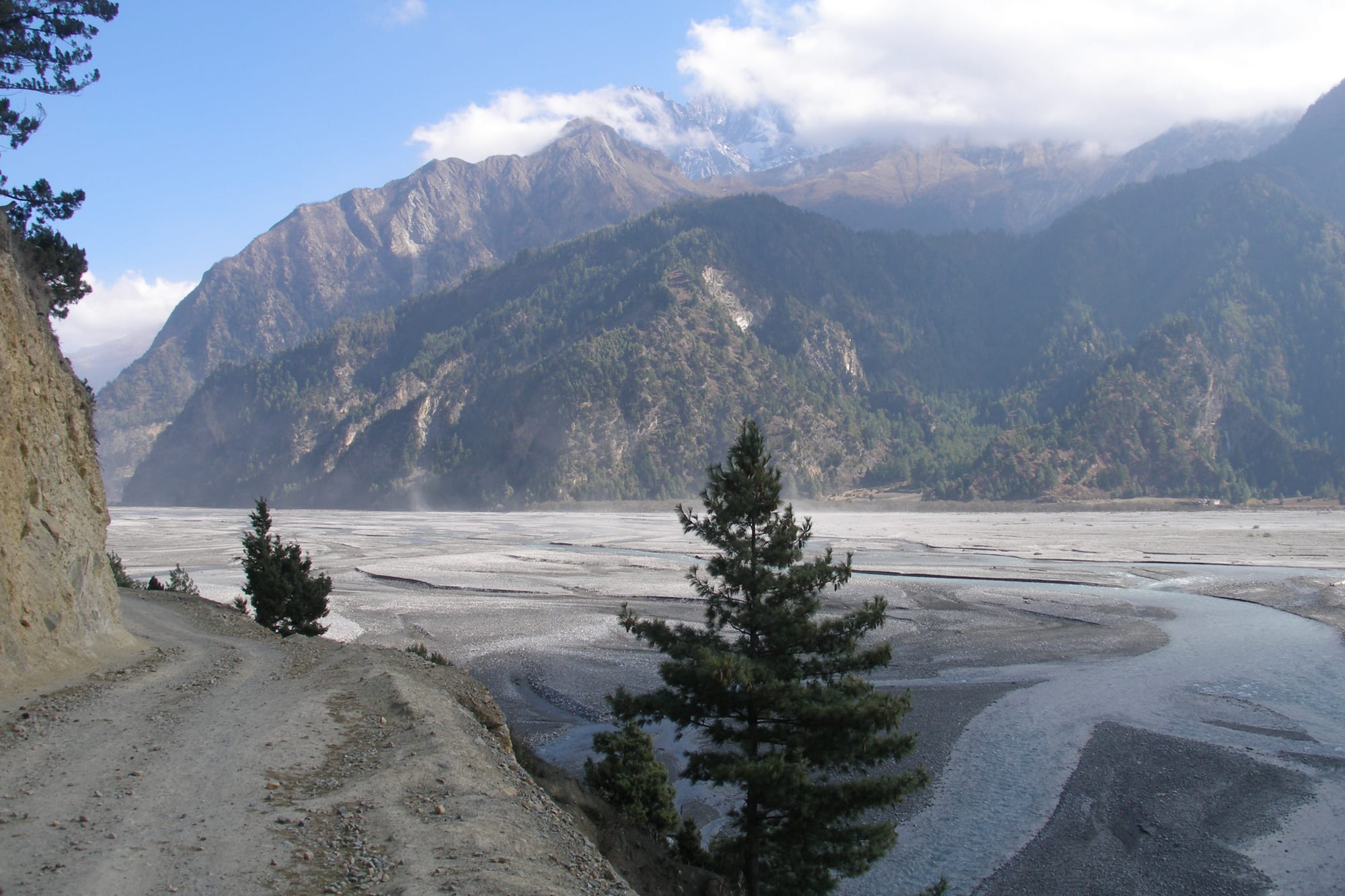 A wide, braided grey riverbed between steep forested mountains, with snow peaks half hidden in cloud beyond