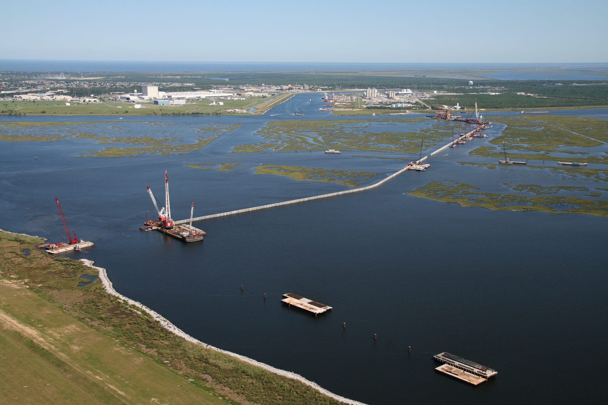 An aerial view of a long concrete wall under construction across open water and marsh, with cranes on barges
