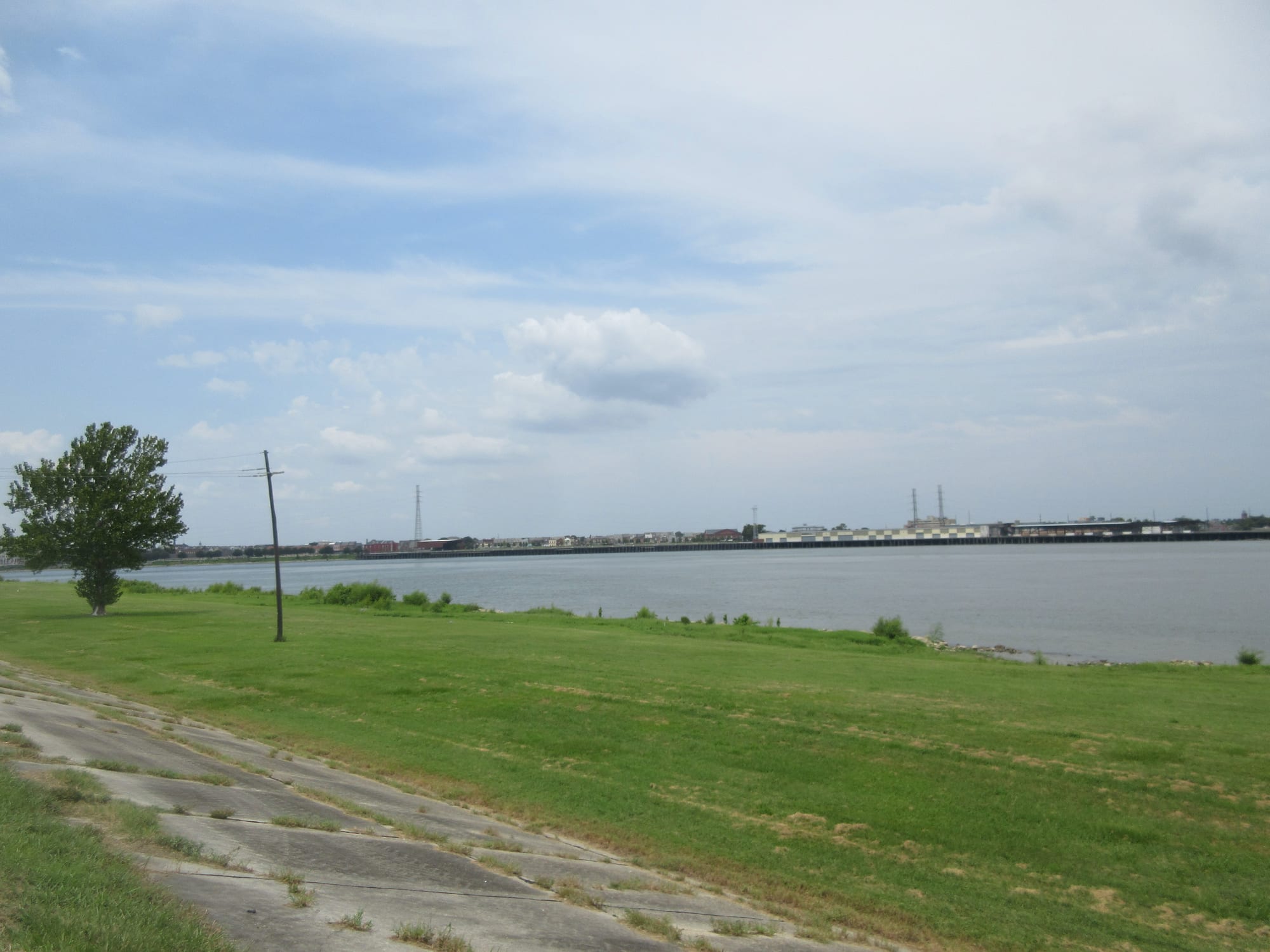 A long grassy embankment with a concrete slope running along the edge of a wide grey river, with industrial buildings on the far bank