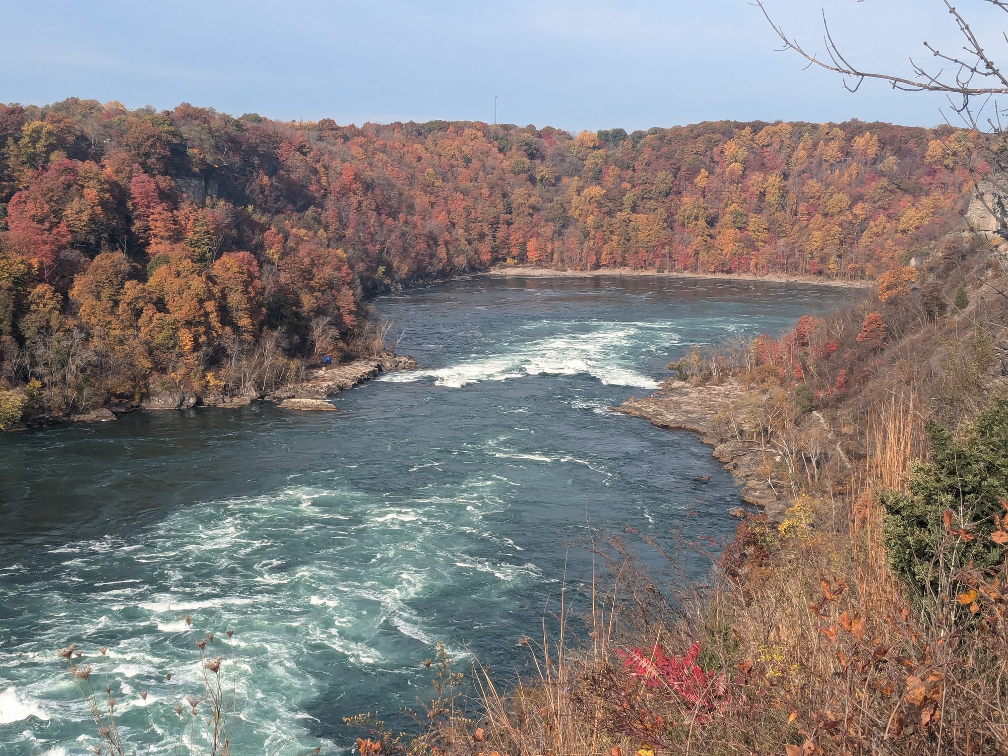 A wide river swirling through a sharp bend between steep banks covered in red and orange autumn trees
