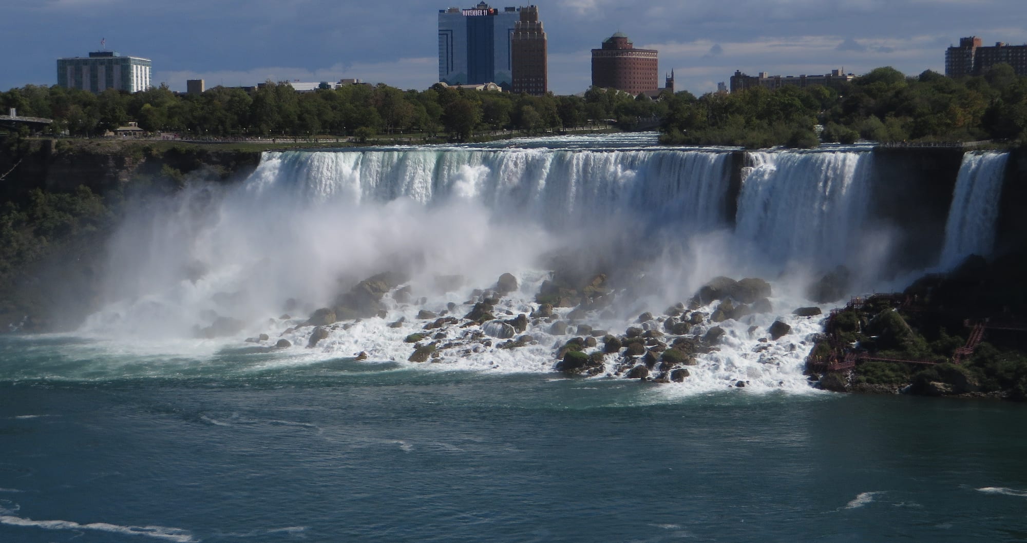 A wide waterfall pouring onto a jumble of large boulders at its base, with spray rising and tall hotel buildings on the bank above