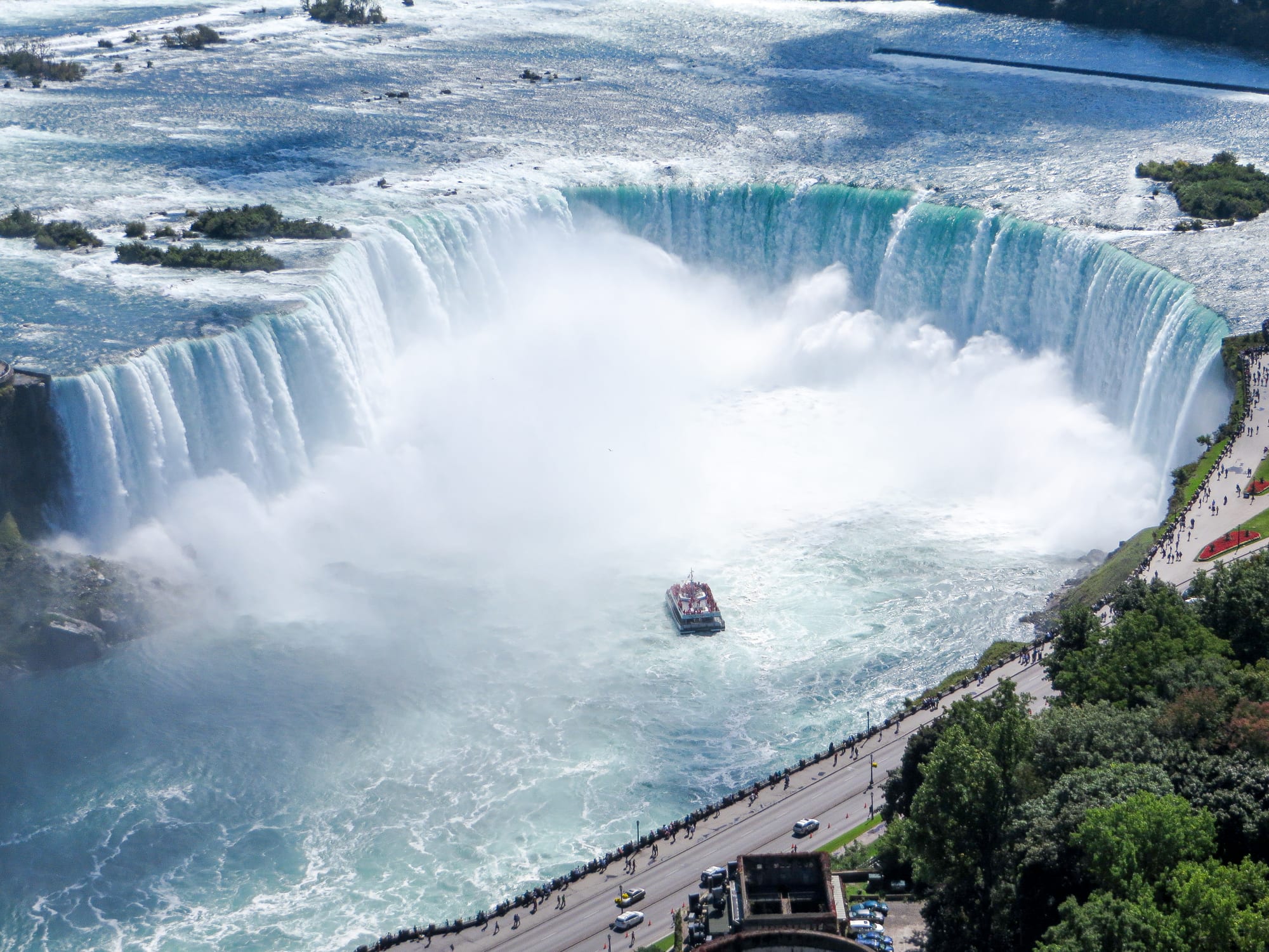 A vast curved waterfall seen from above, with clouds of spray rising from the basin, a small tour boat near its foot and a riverside road below
