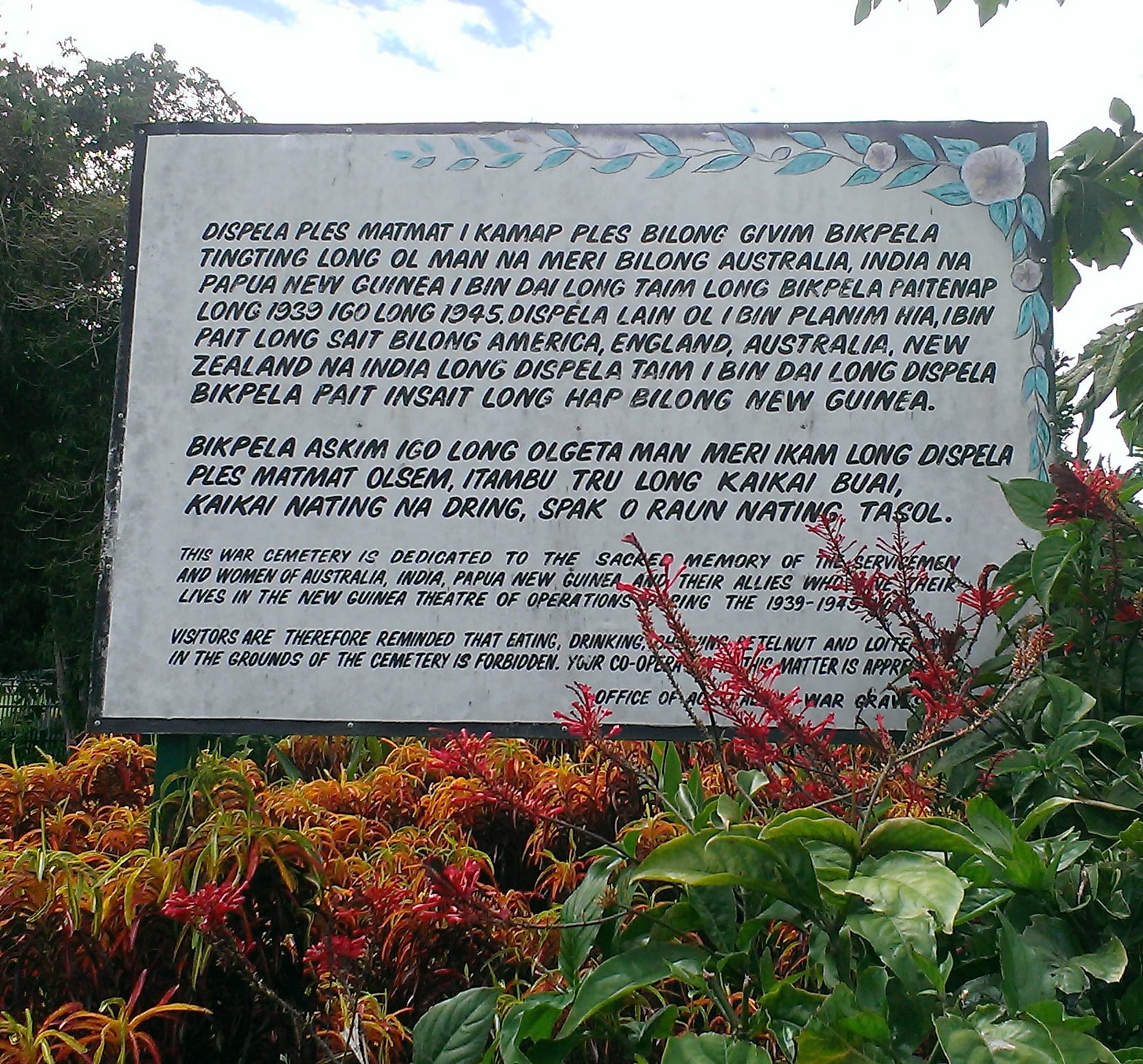 A large painted sign with paragraphs in Tok Pisin above smaller text in English, framed by red flowers