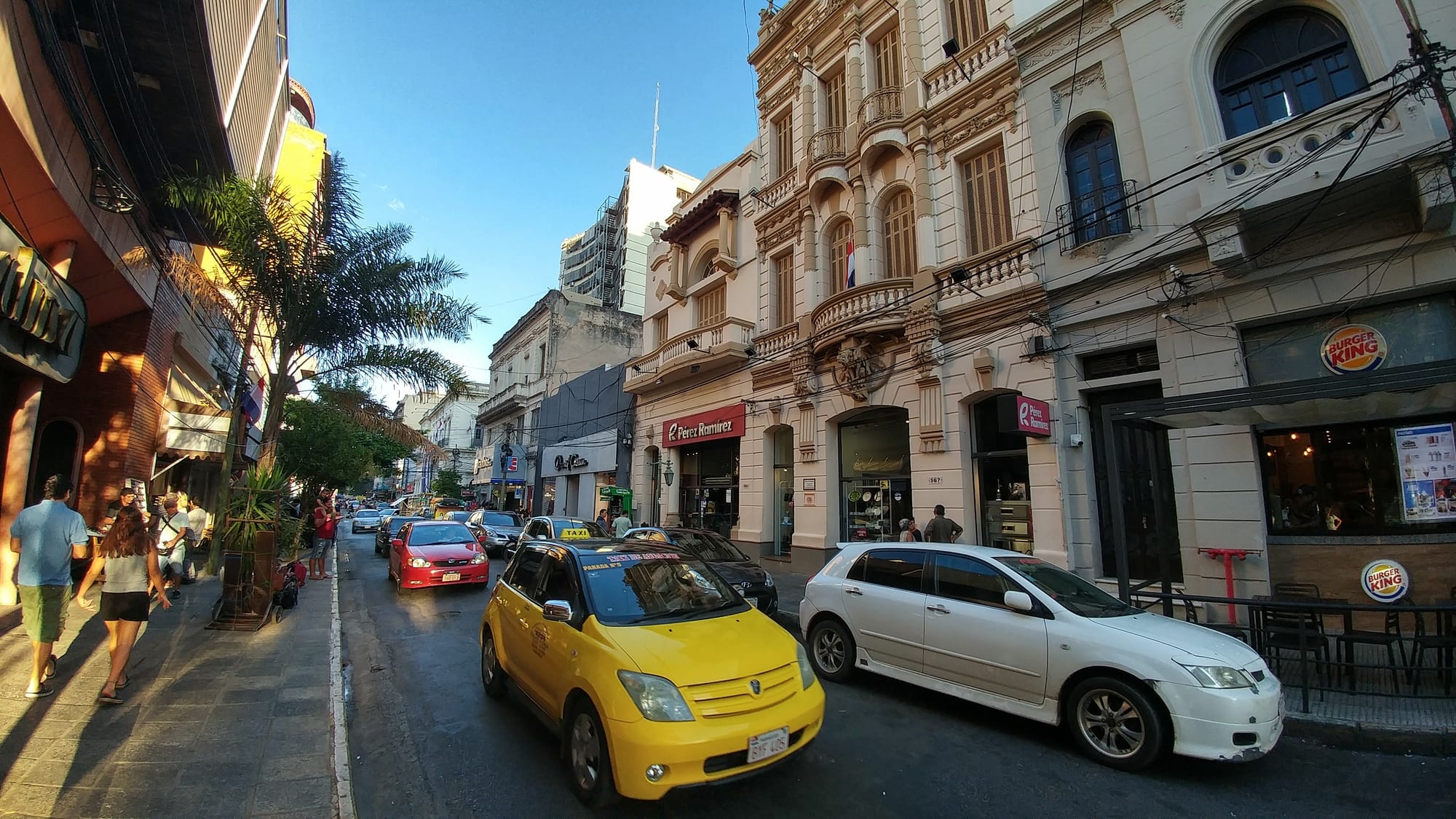 A busy downtown street lined with ornate cream-coloured colonial buildings, with a yellow taxi and other cars in the road and people on the pavement