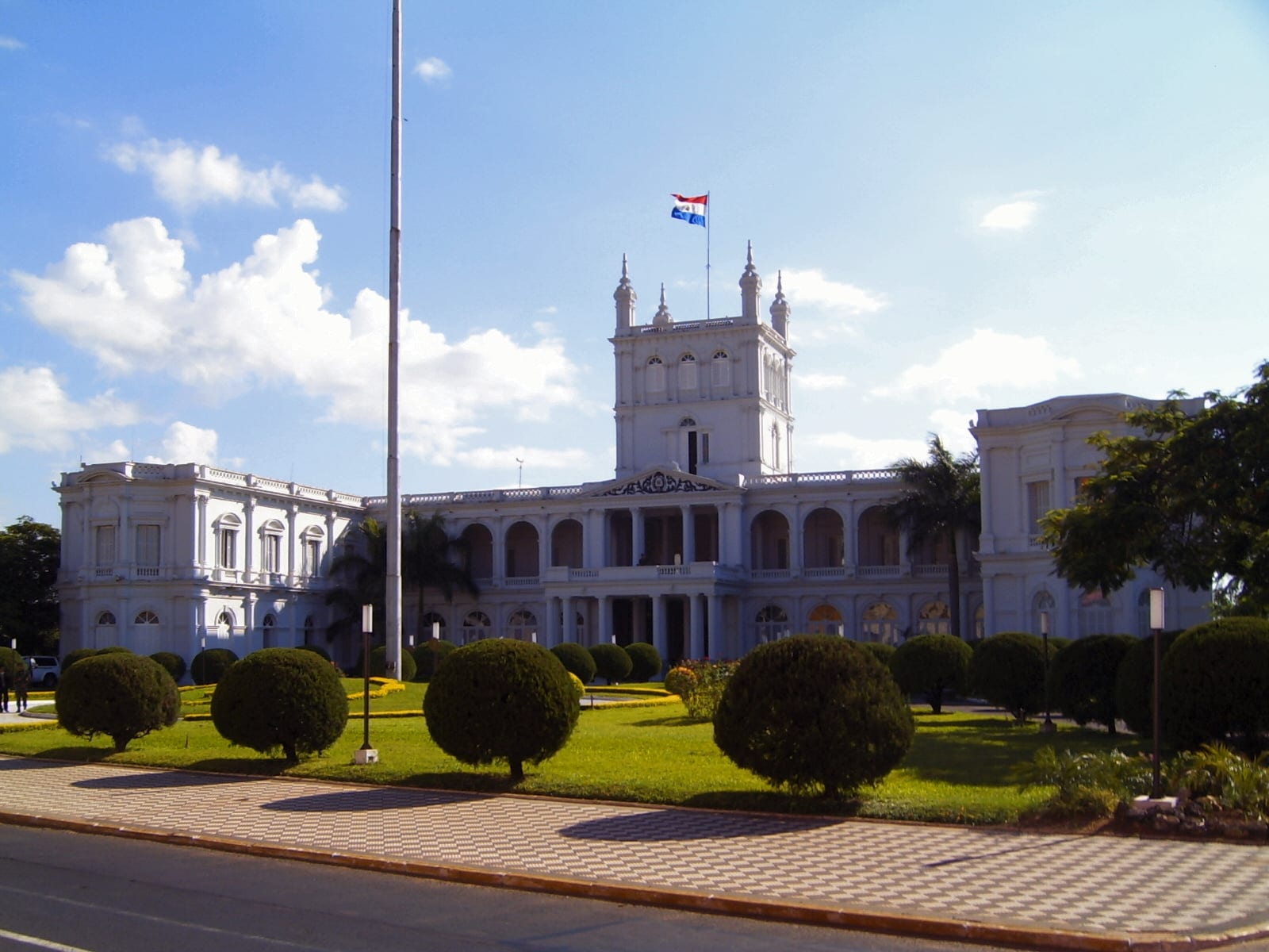 A white neoclassical palace with arched galleries and a central tower flying the red, white and blue Paraguayan flag, behind rounded topiary bushes