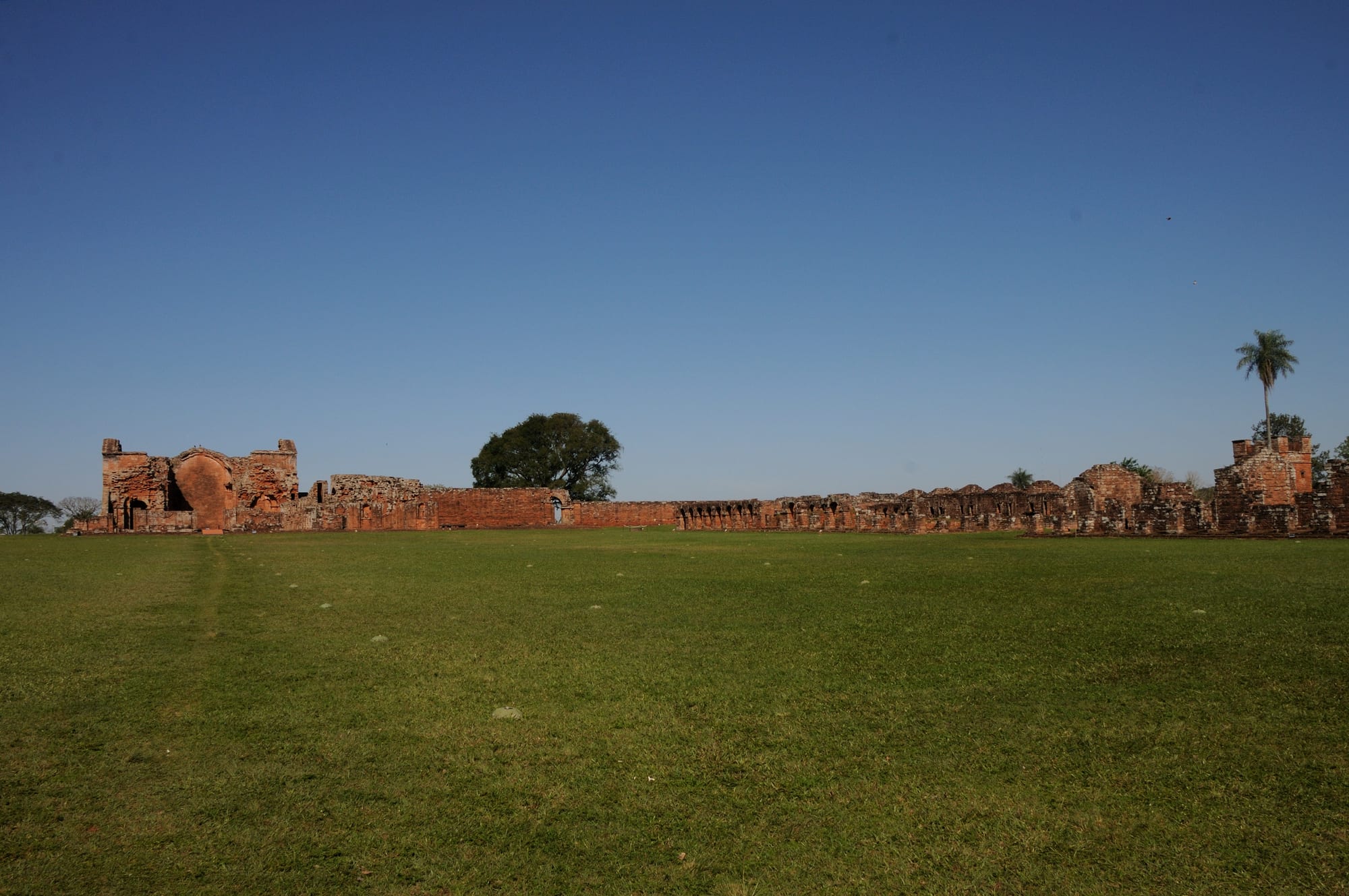 A long line of roofless red sandstone ruins and a broken church facade beyond a wide, flat lawn under a clear blue sky