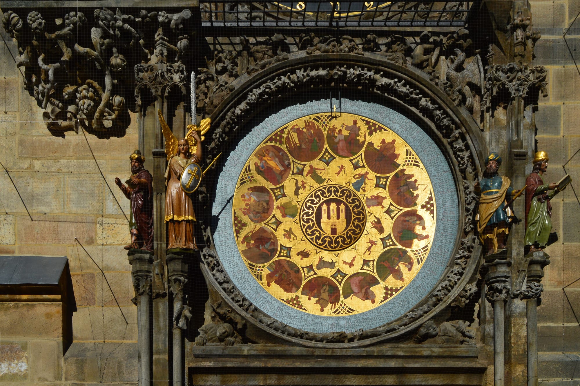 A round golden calendar plate painted with medallions of country scenes and a small castle emblem at the centre, flanked by carved and painted statues