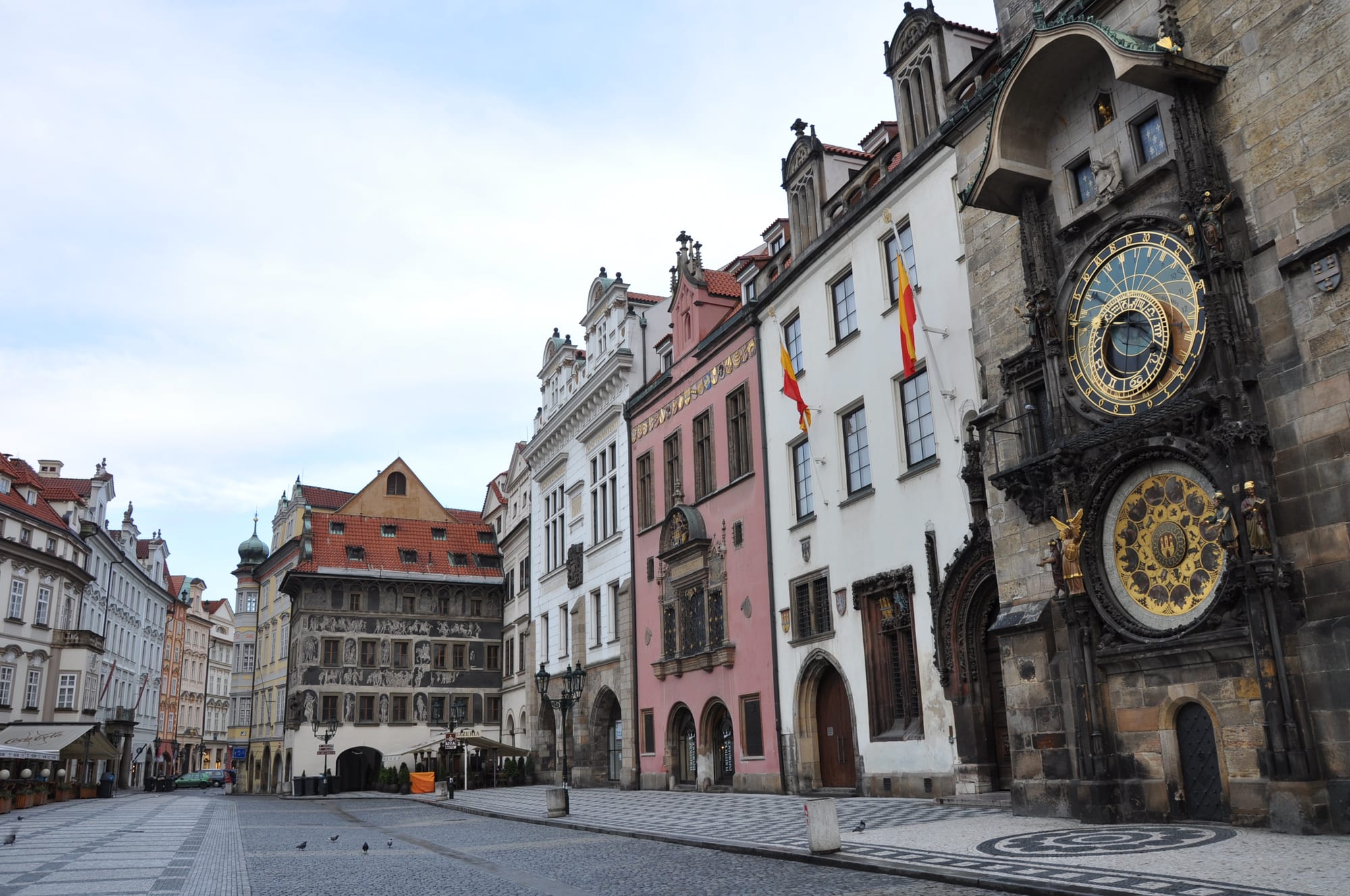 A quiet cobbled square lined with pastel-coloured houses, with a stone tower on the right bearing a gold astronomical dial above a round calendar plate
