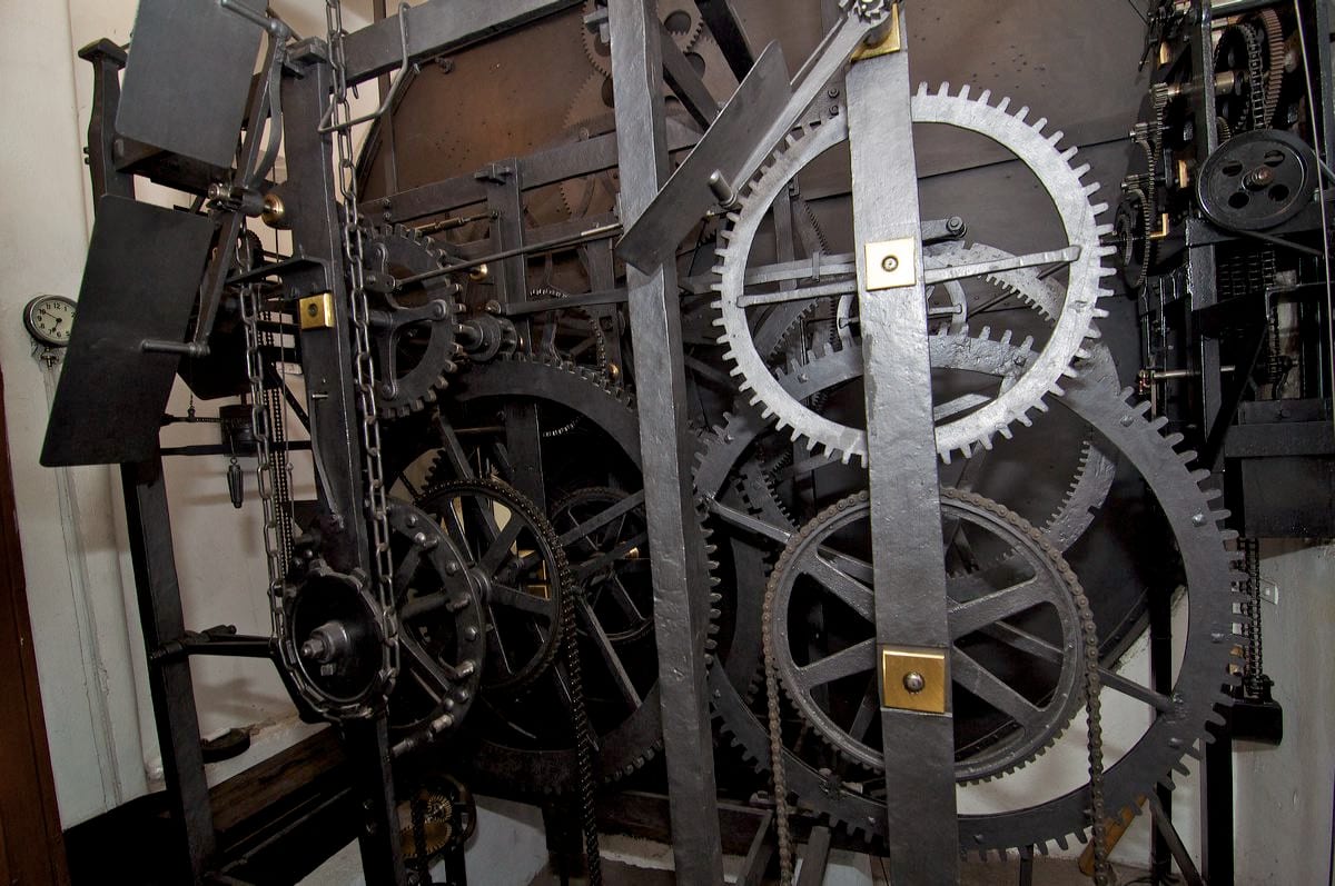 Large black iron gear wheels, chains and frames of an old clock mechanism inside a white-walled room