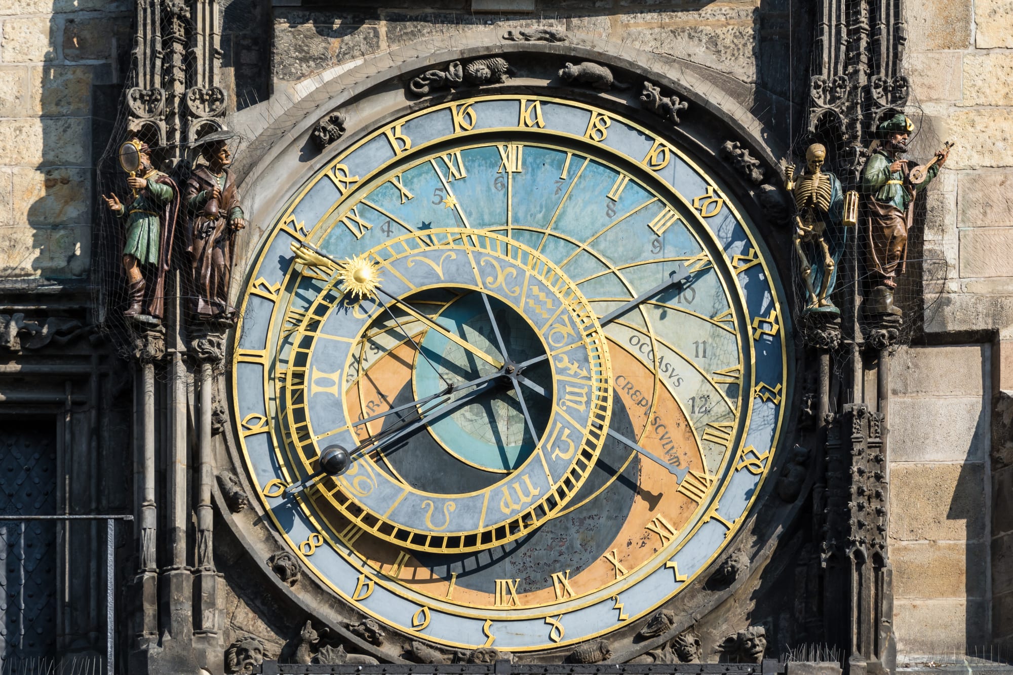 A large round clock face in blue, gold, black and orange with a gilded sun on a long hand, flanked by small carved figures including a skeleton
