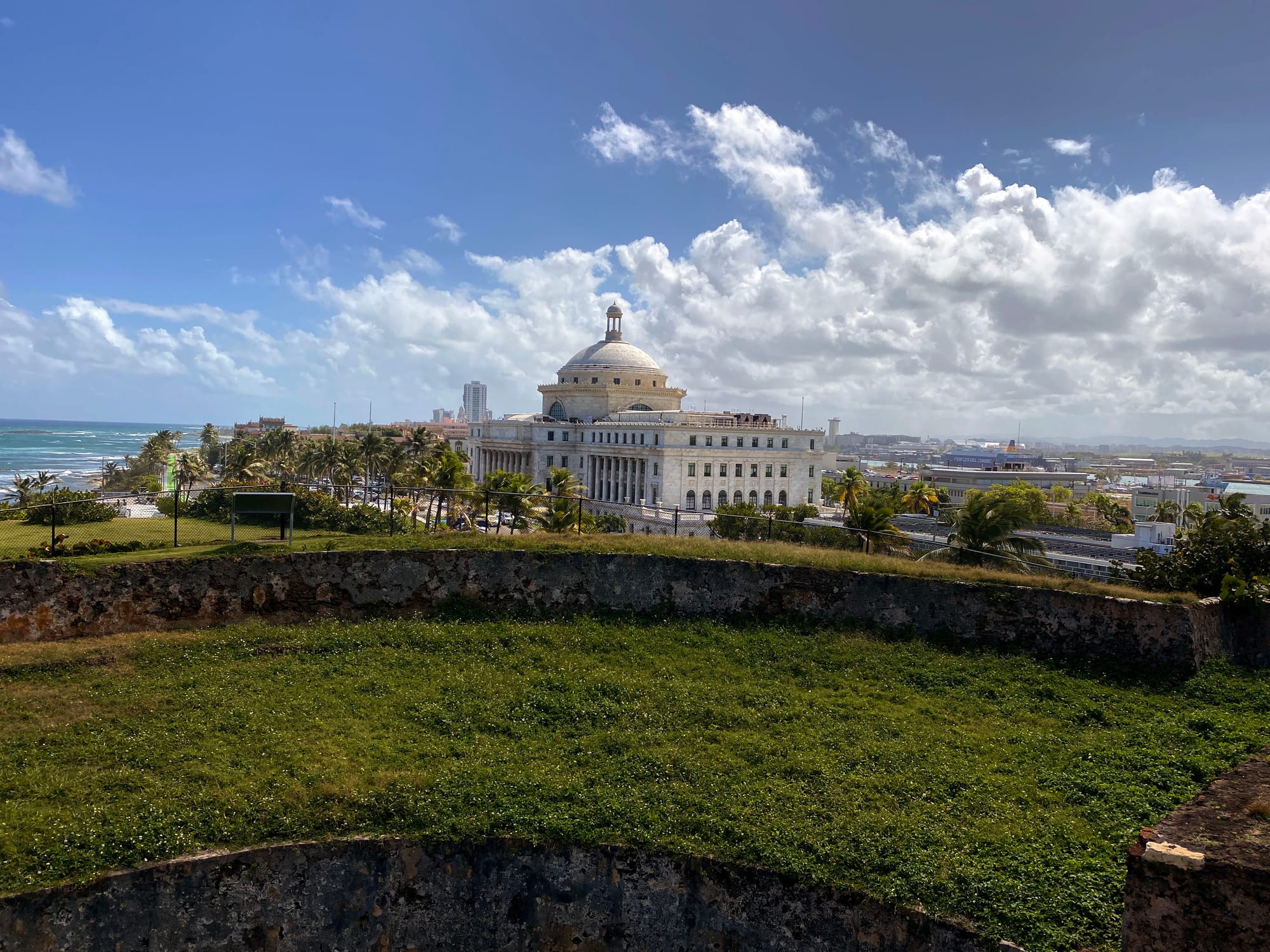 A white domed neoclassical building among palm trees beside the ocean, seen across the grassy walls of an old fort under a blue sky