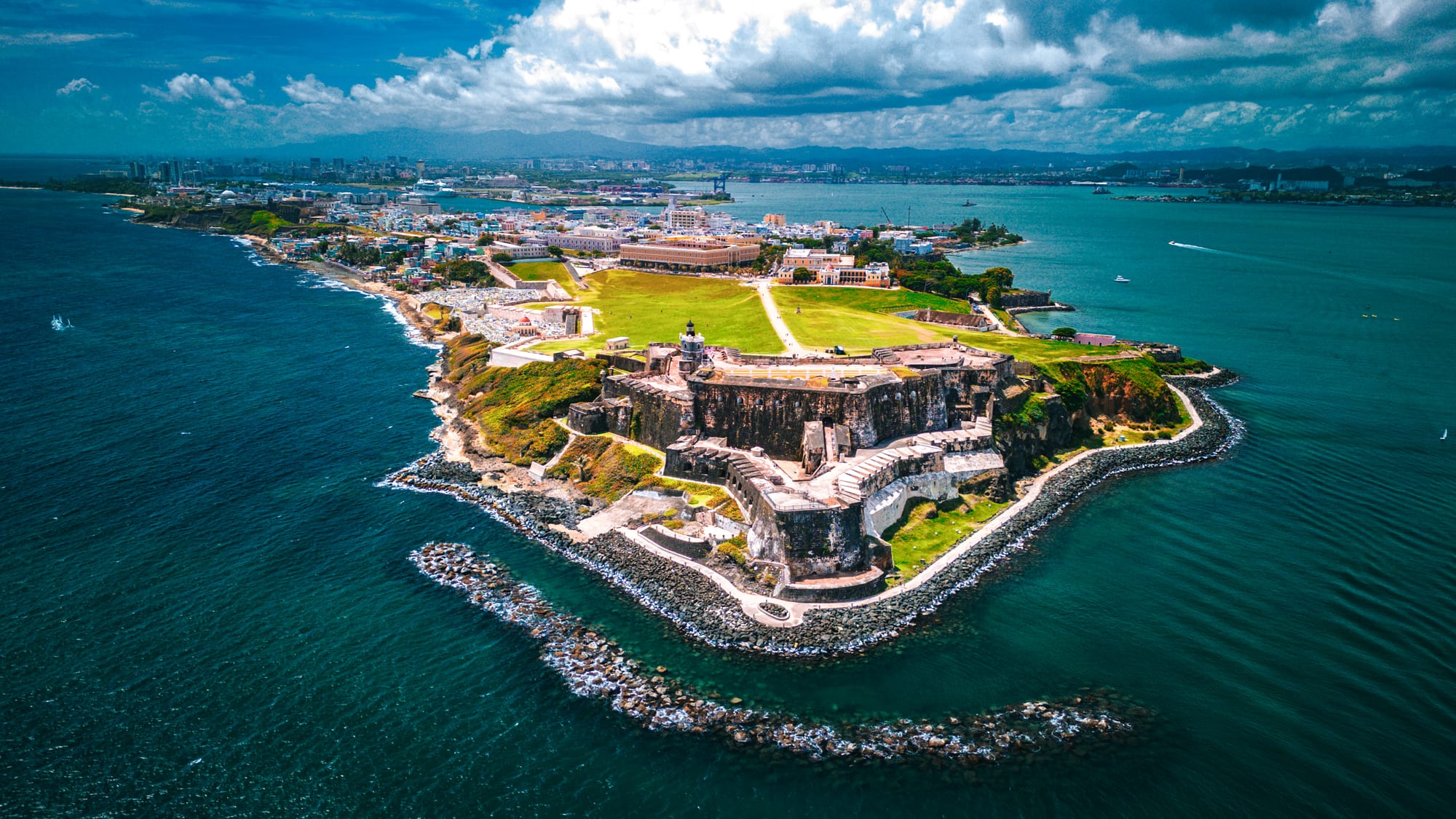 An aerial view of a large stone fortress on a rocky headland surrounded by deep blue sea, with the old city and green lawns behind it