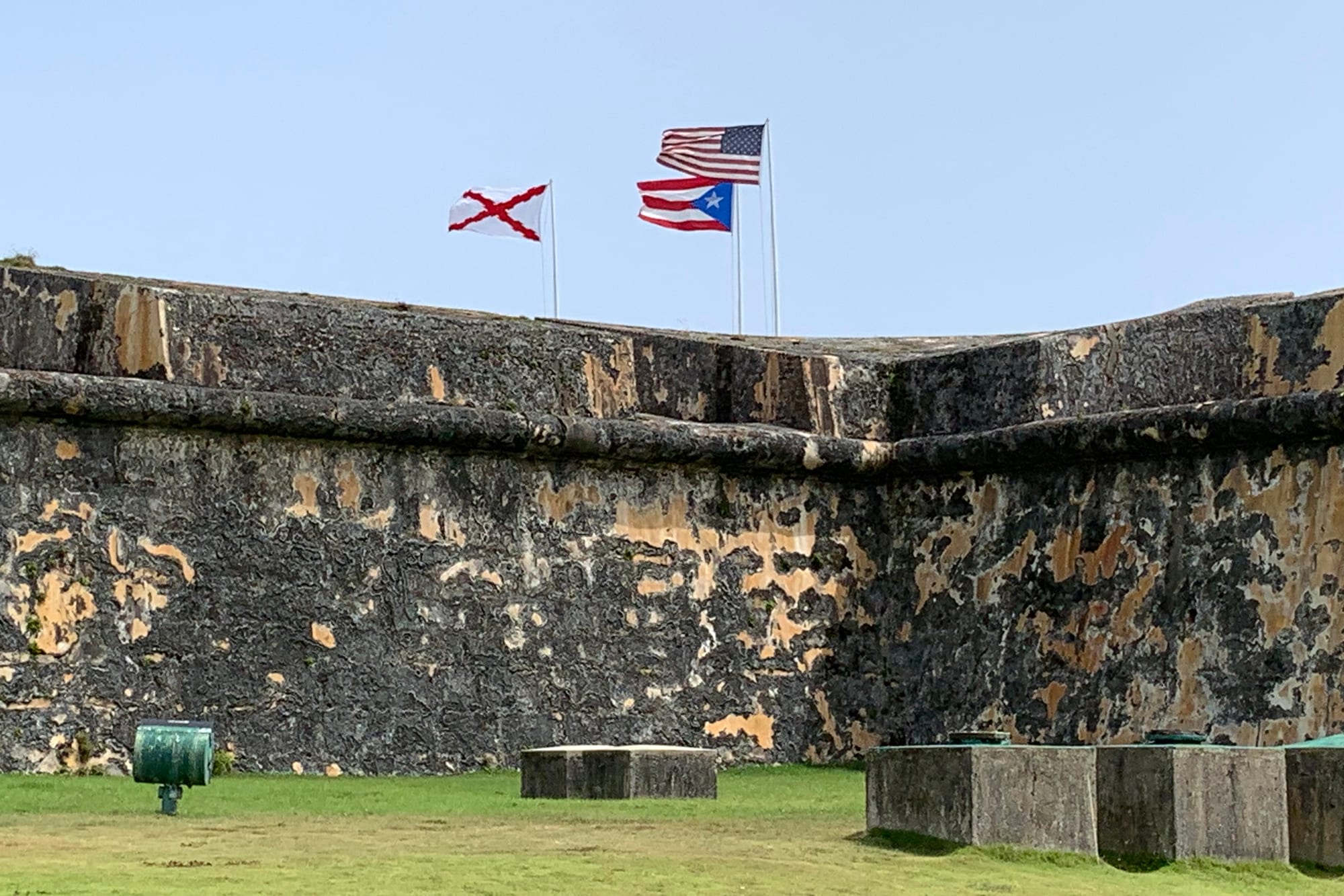 Three flags flying above the weathered stone wall of an old fort: the Stars and Stripes, the Puerto Rican flag and a white flag with a red diagonal cross