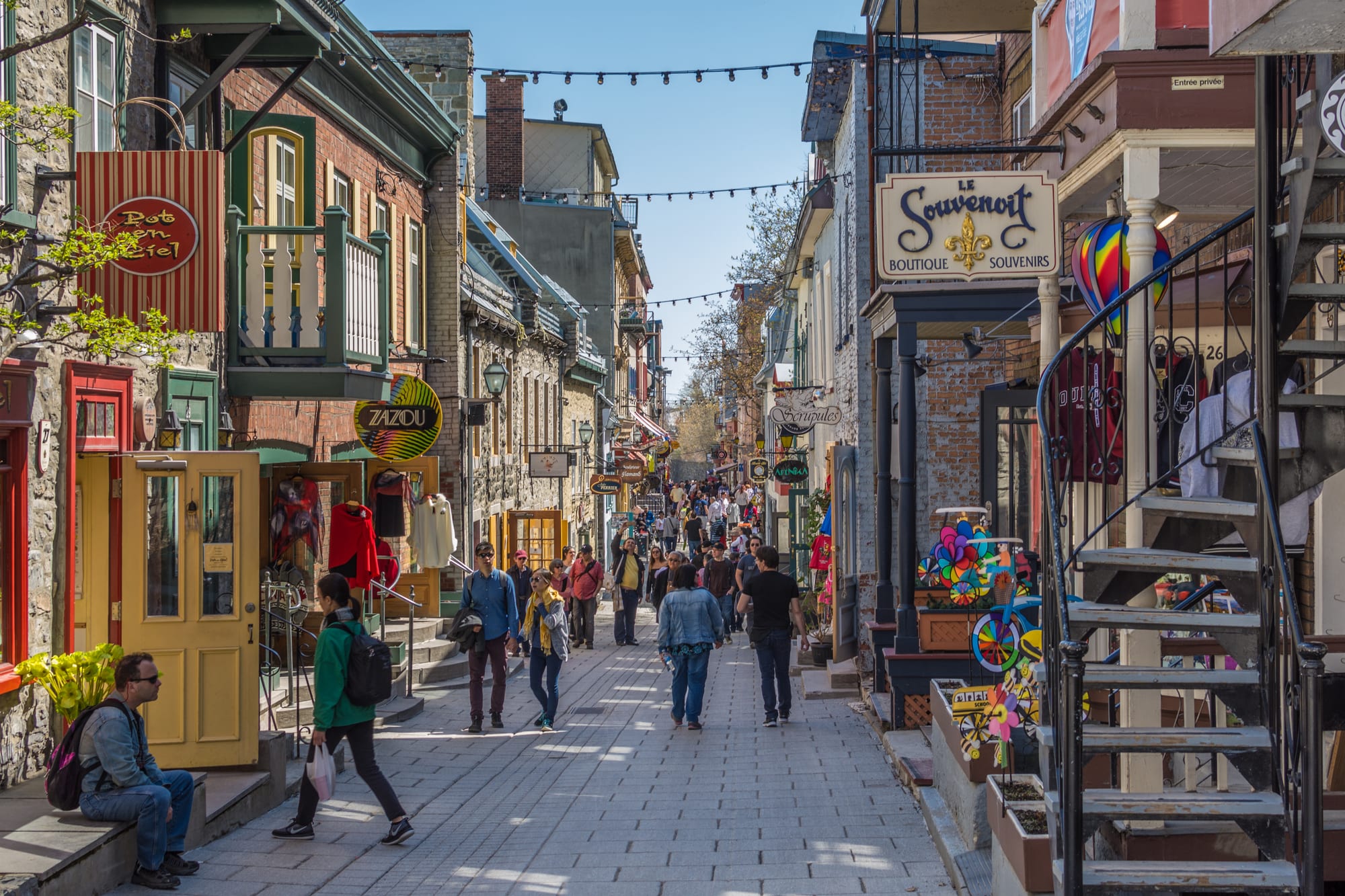 A busy pedestrian street of old stone and brick houses with shop signs in French and strings of lights overhead