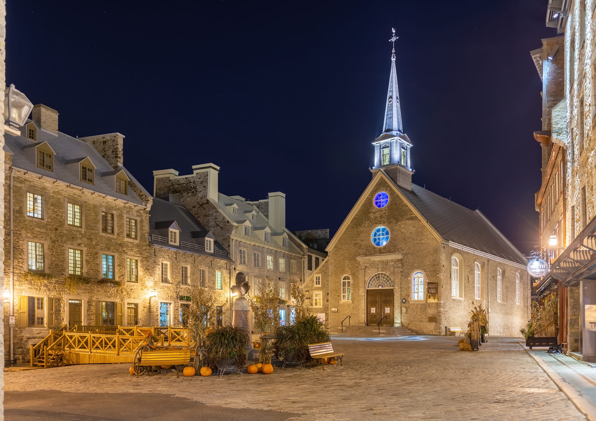 A small cobbled square at night, lined with tall grey stone houses with steep roofs and dormer windows, and a stone church with a lit spire