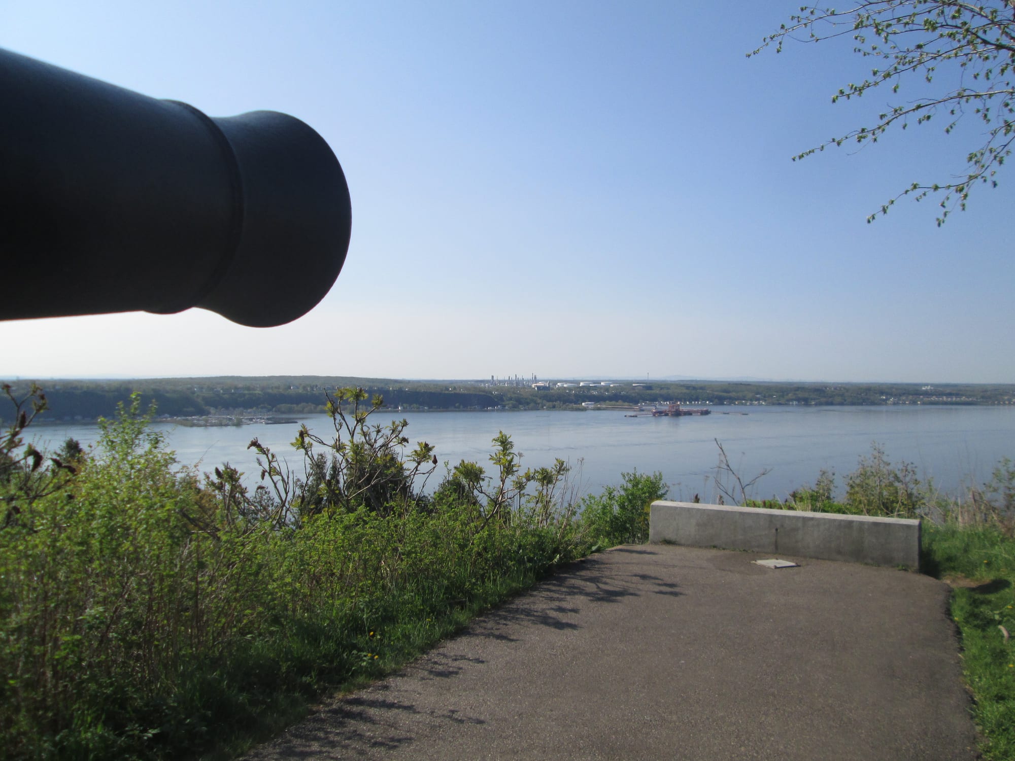 The muzzle of an old black cannon pointing over green bushes towards a wide river and a low wooded far shore