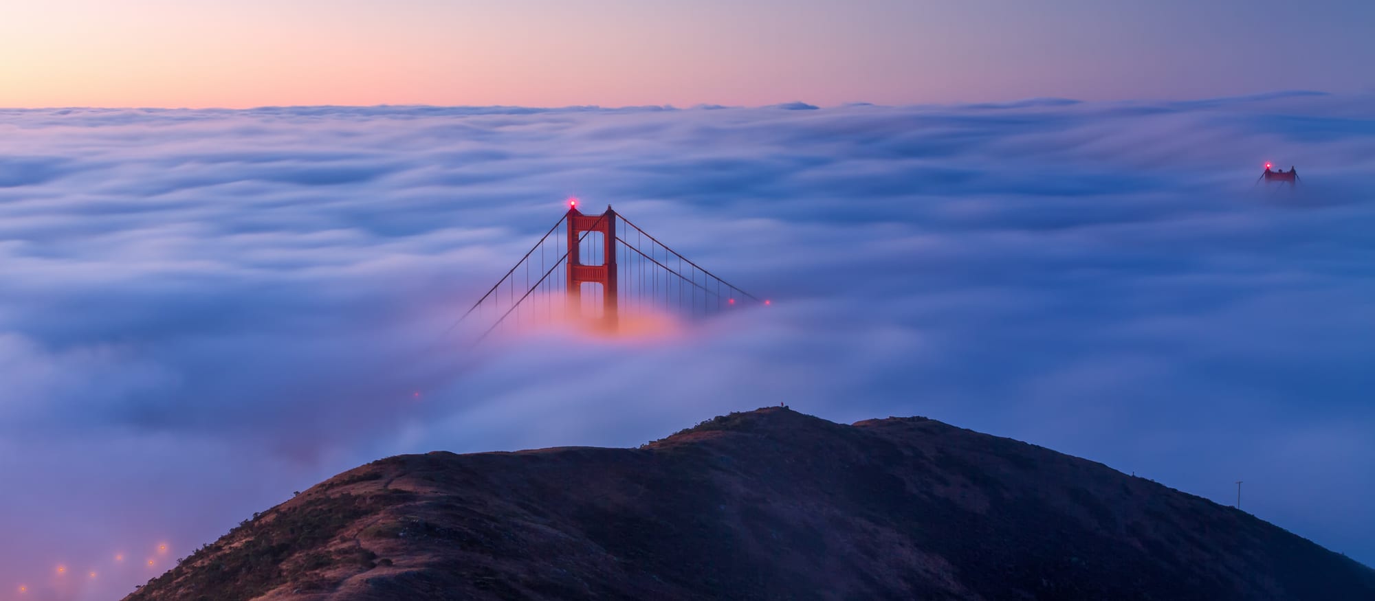 The red tower of a suspension bridge rising out of a thick white sea of fog at dawn, seen from a dark hilltop, with a pink sky above