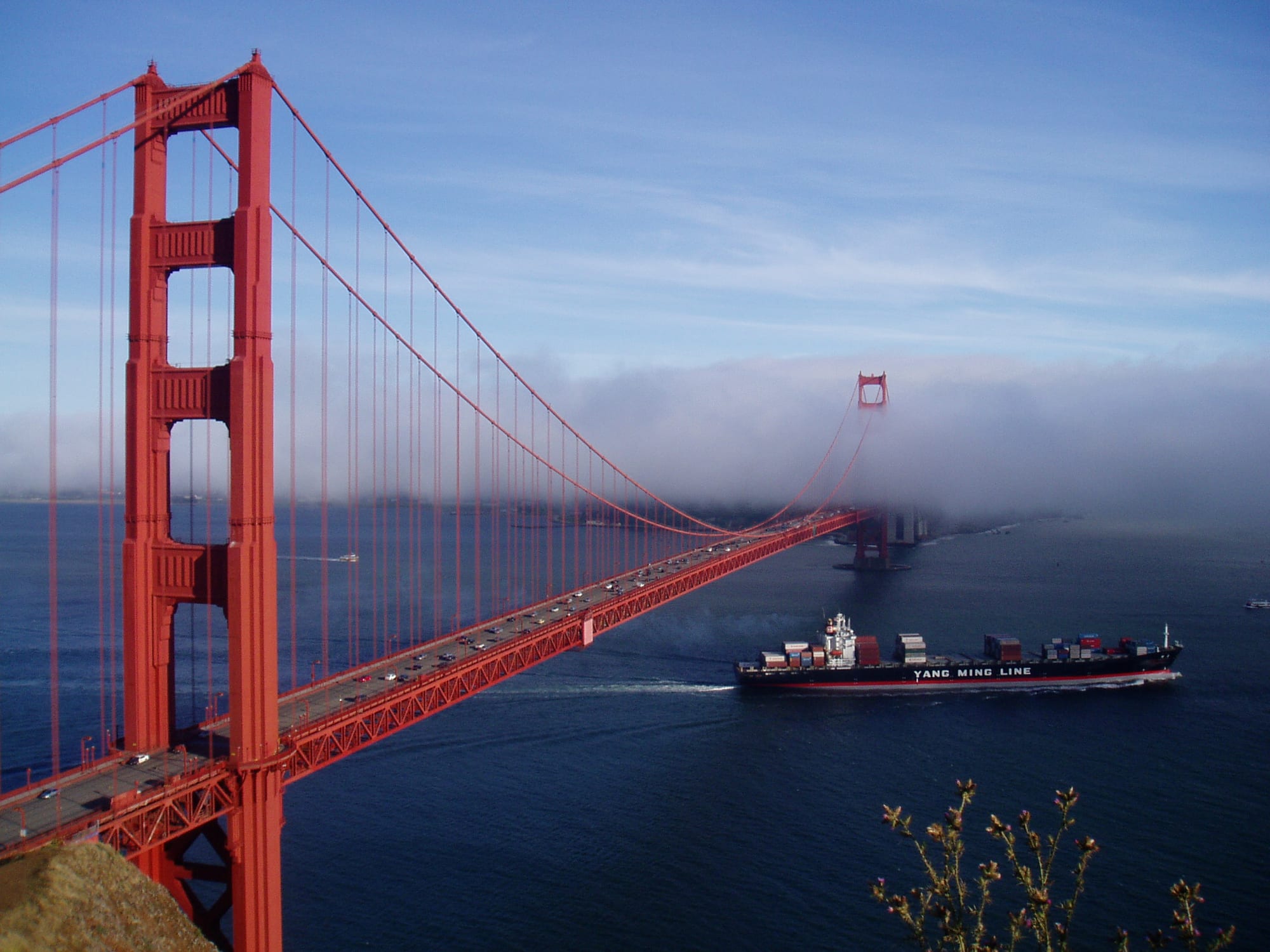 A red suspension bridge crossing a blue strait in sunshine, with a bank of fog swallowing its far tower and a container ship passing beneath