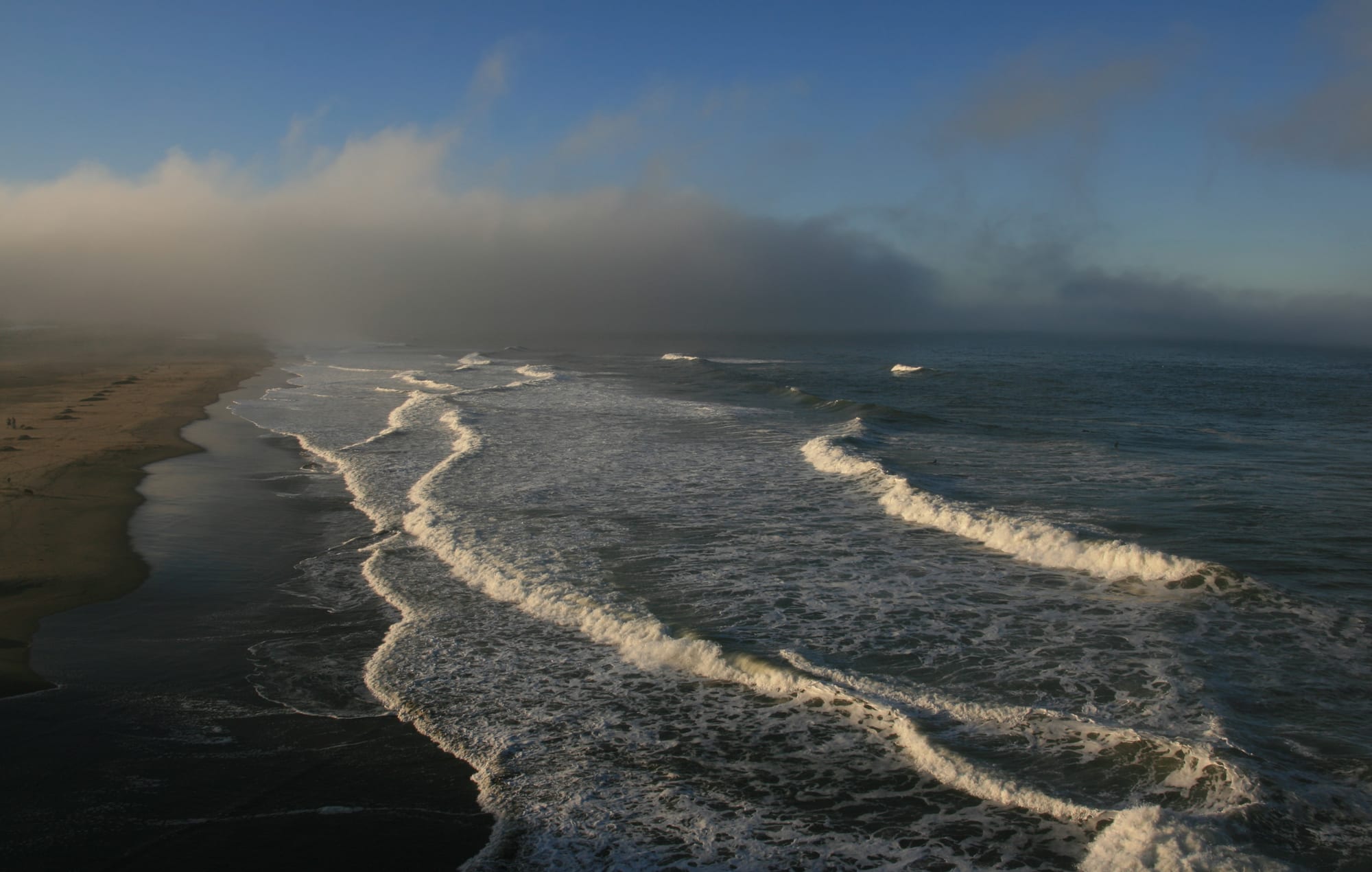 Breaking waves rolling onto a long sandy beach, with a grey bank of fog lying low over the sea and shore under a blue sky