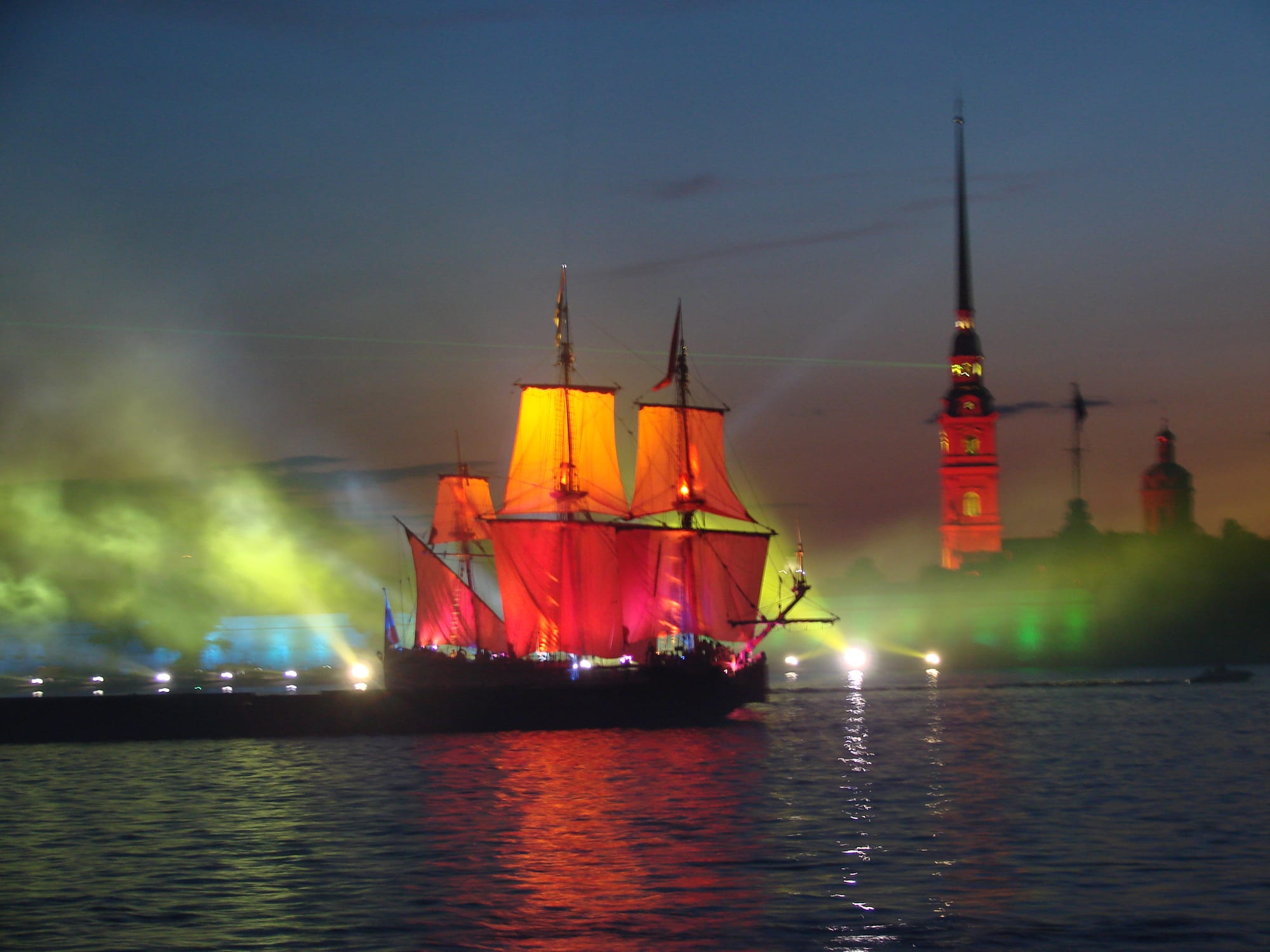A three-masted sailing ship with glowing red sails on a river at night, lit by green and yellow searchlights, with a floodlit spire behind