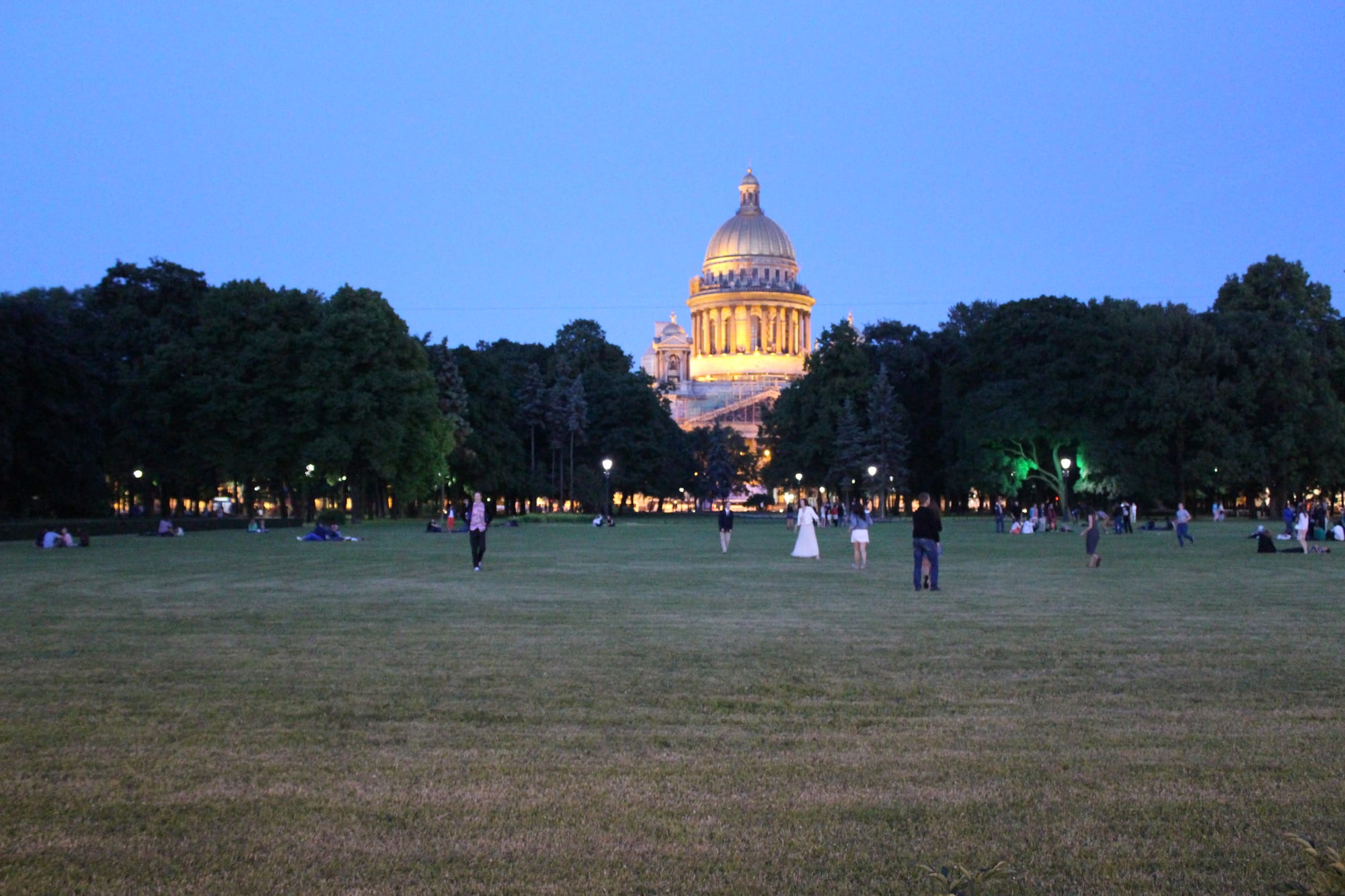 People walking and sitting on a large lawn at dusk, with a floodlit golden-domed cathedral rising above dark trees under a blue sky