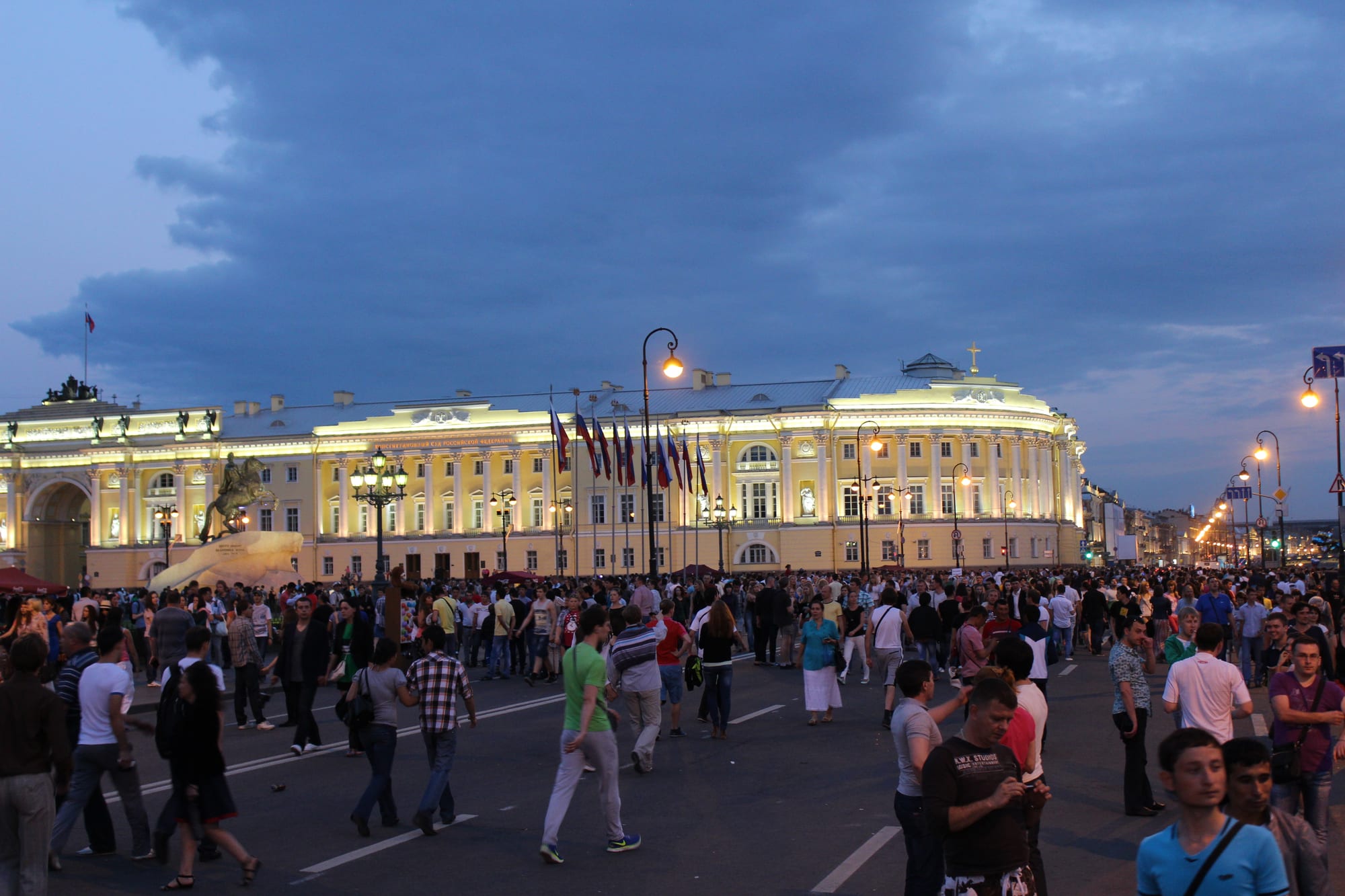 Crowds walking on a wide street in front of a long floodlit yellow and white classical building under a deep blue evening sky, with an equestrian statue on the left
