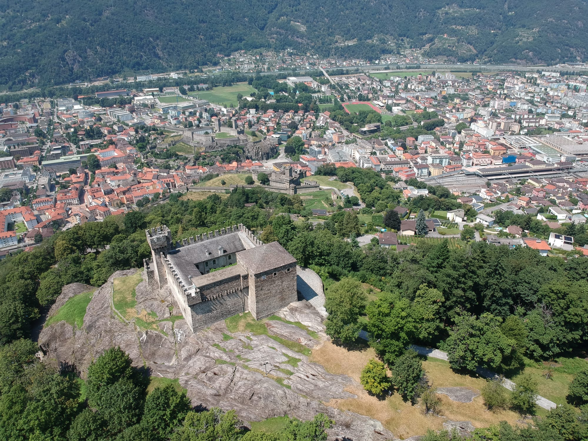 A stone castle on a rocky wooded hill, seen from above, with a town of red-roofed houses and a second walled castle in the valley below