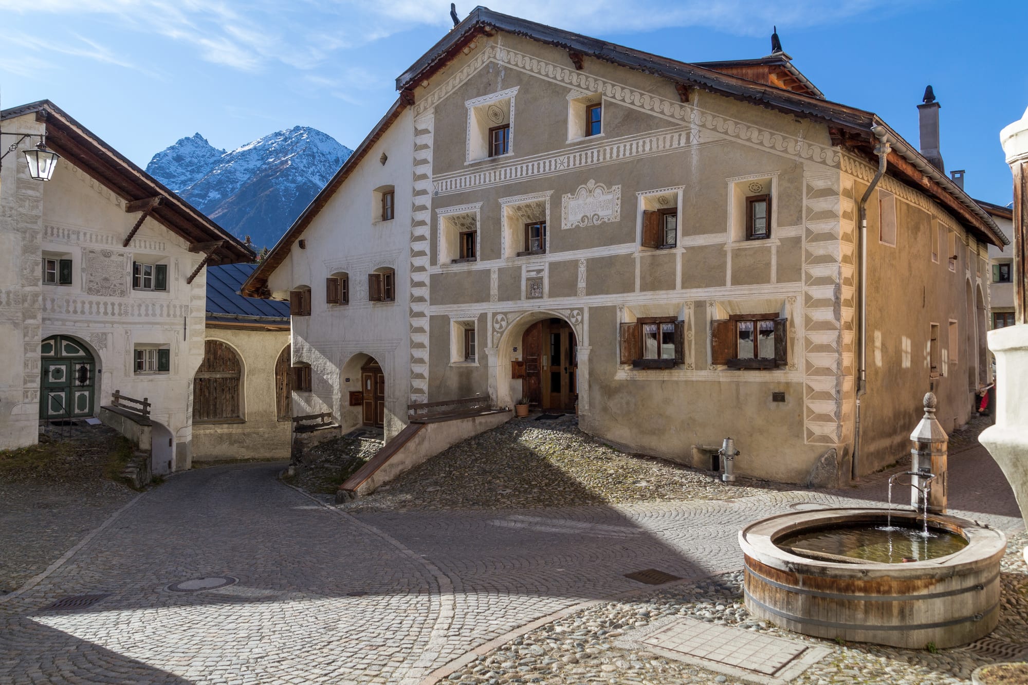 A large village house with grey and white geometric plaster decoration, beside a wooden water trough and a cobbled square, with a snowy peak behind