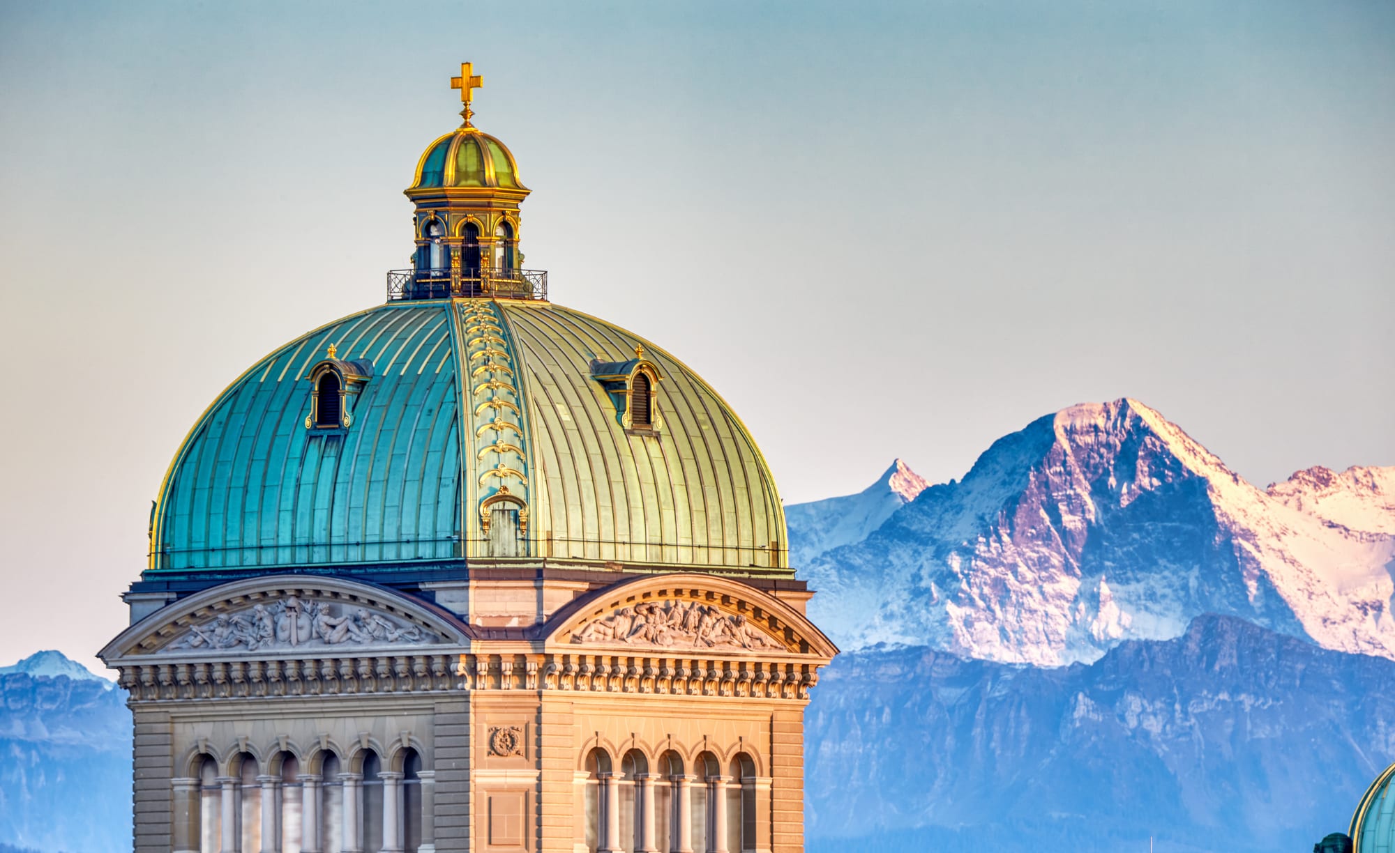 The green copper dome of a large stone parliament building, topped with a gilded cross, in front of a snow-covered mountain lit pink by the evening sun