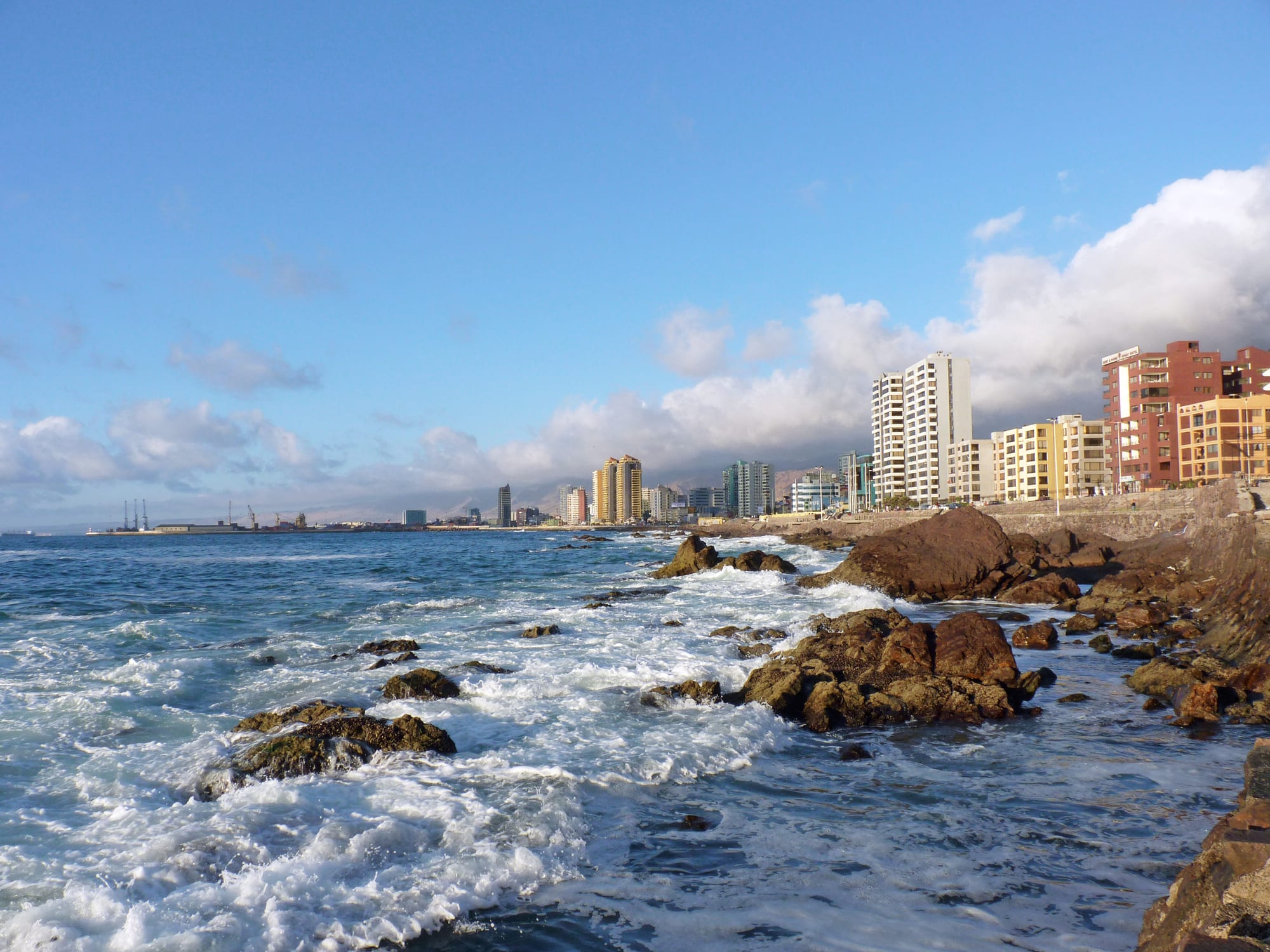 Waves breaking on brown rocks in the foreground, with a line of apartment towers along the shore and a bank of cloud over hills behind
