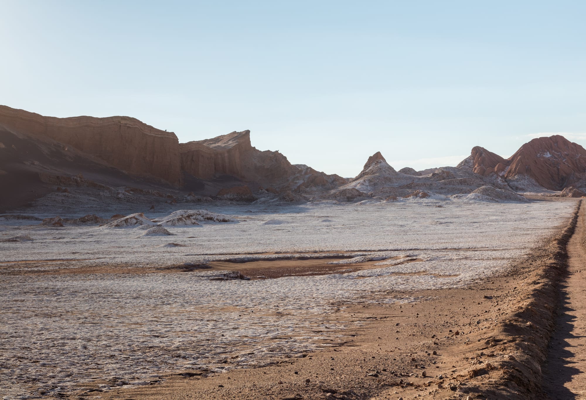 A pale, crusted desert floor with no plants, ringed by eroded brown and pink rock ridges under a clear sky