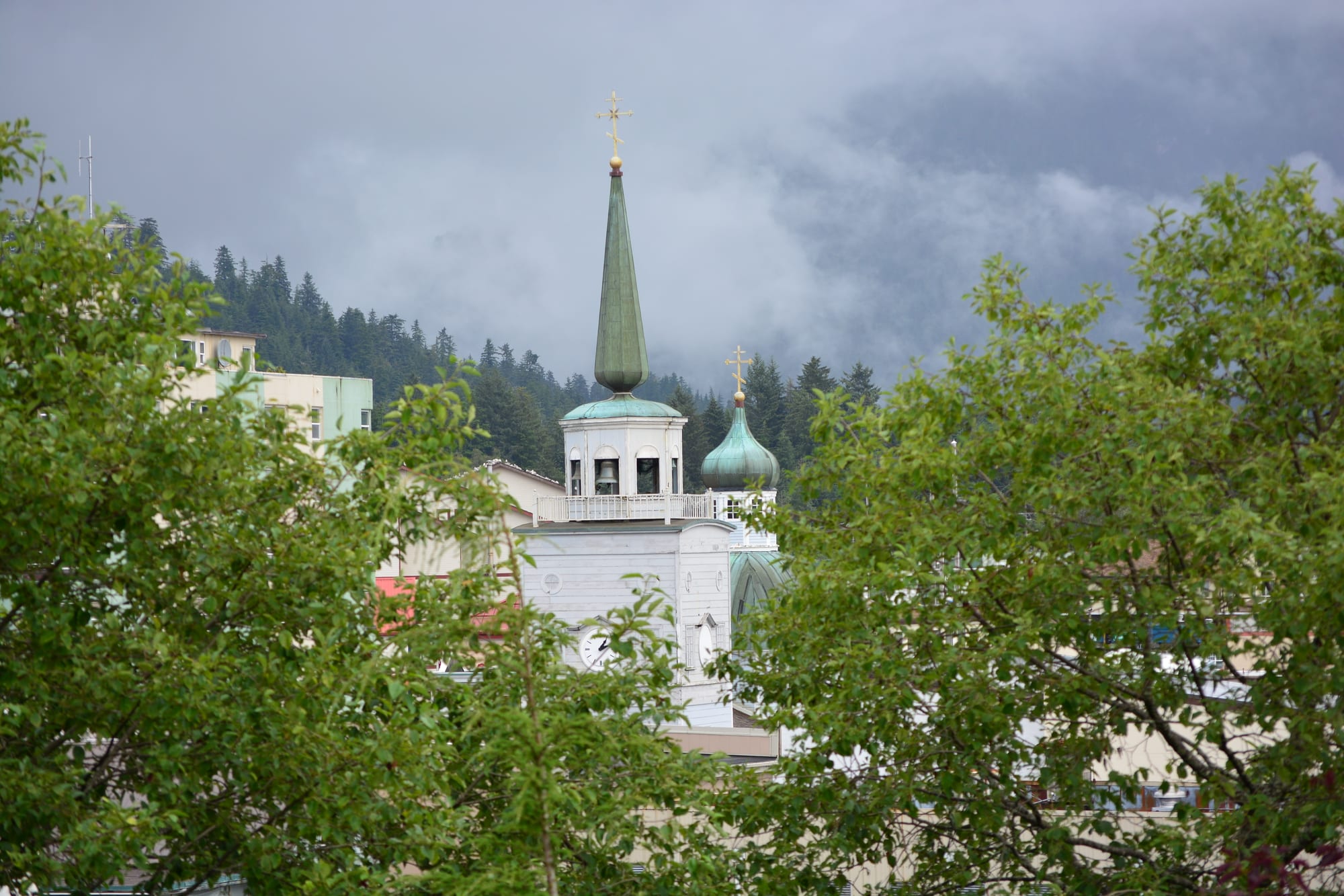 A white church tower with a green spire and onion dome rising among trees below misty forested hills