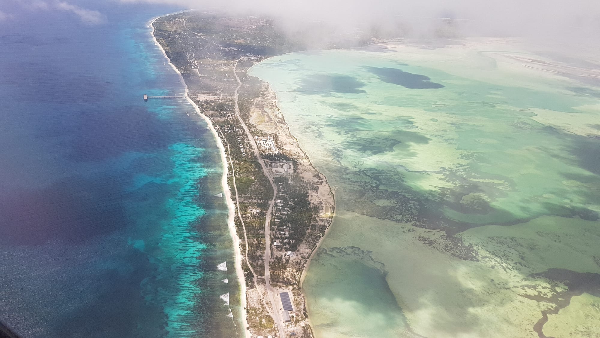 An aerial view of a narrow strip of island with a town and a road, between deep blue ocean and a pale green lagoon