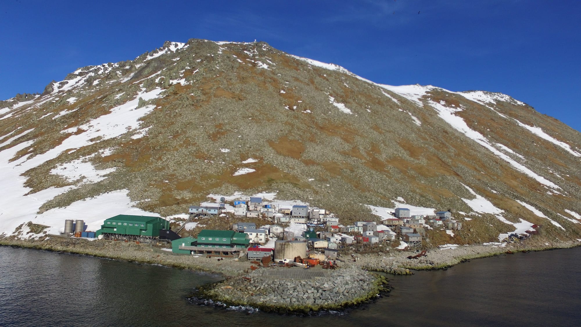 Small houses and green-roofed buildings at the foot of a steep, snow-streaked rocky island above dark water