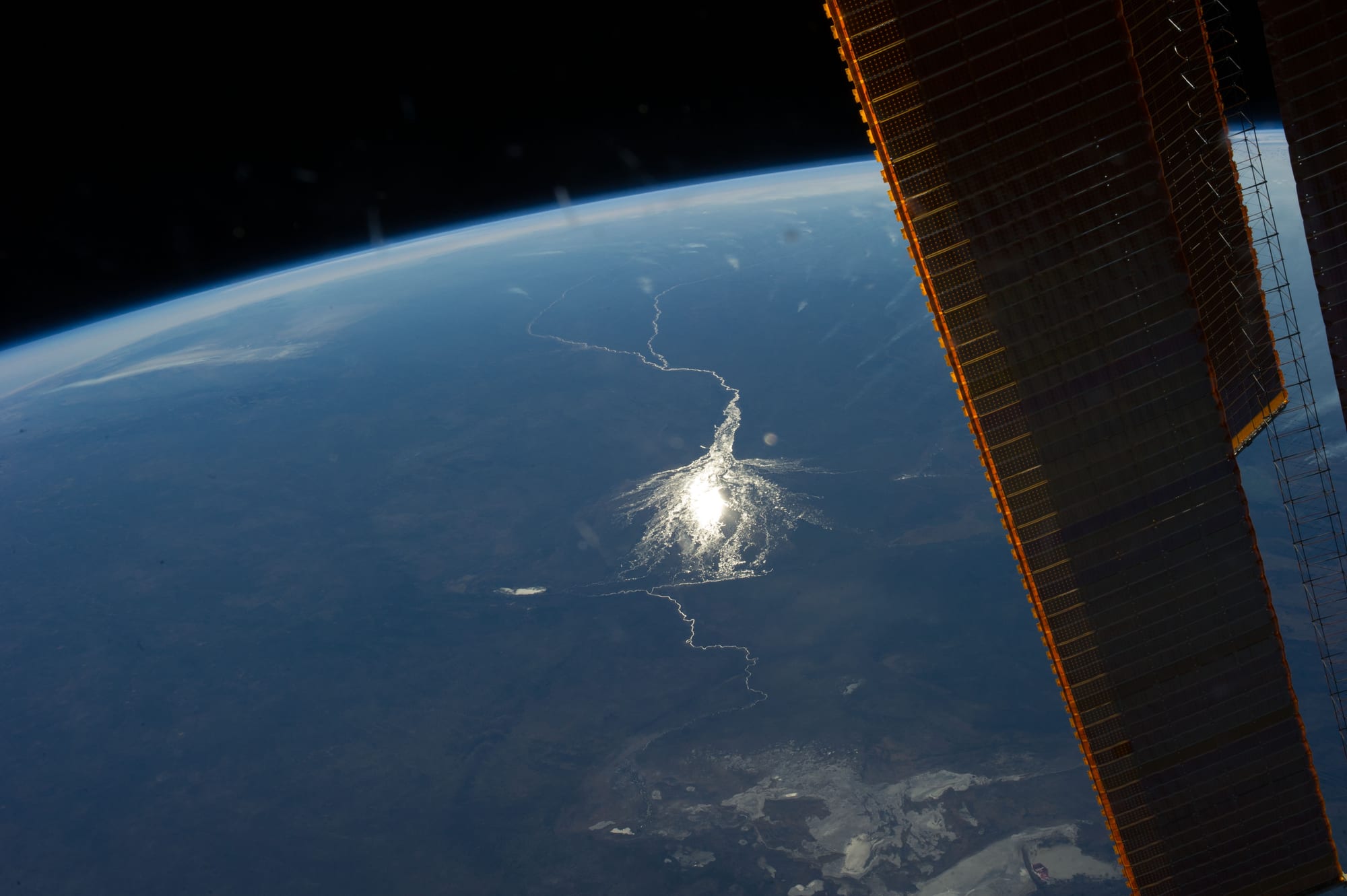 A thin silver river seen from space spreading into a bright, fan-shaped patch of water on a dark brown plain, with part of a spacecraft's solar panel on the right