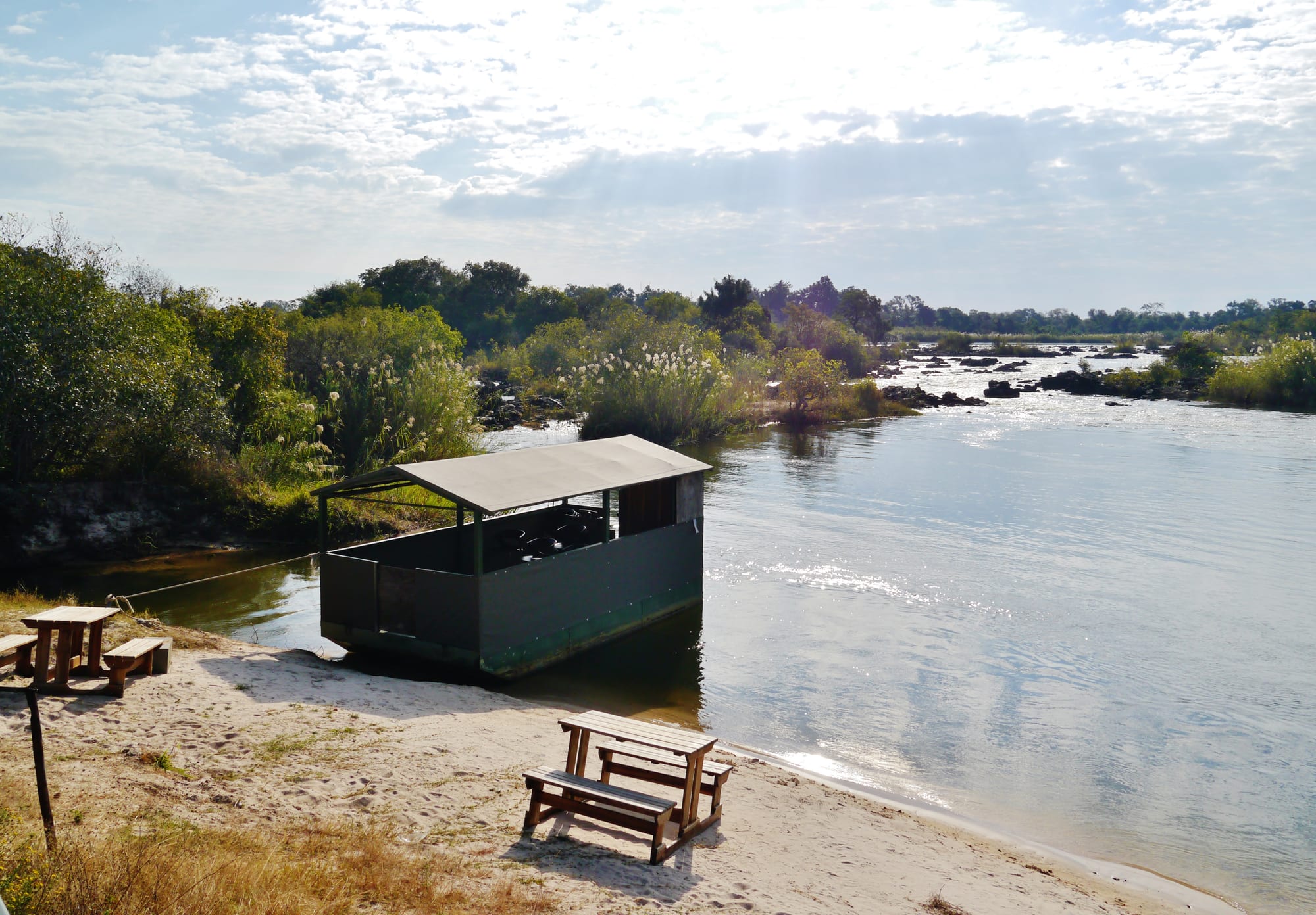 A wide, calm river with rocky rapids in the distance, a covered wooden platform moored at a sandy bank and picnic benches in the foreground