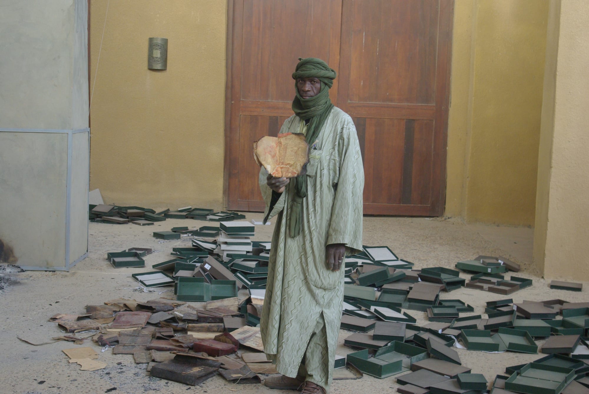 A man in a long robe and green turban holds up a scorched manuscript page in a room strewn with empty green boxes and burnt papers