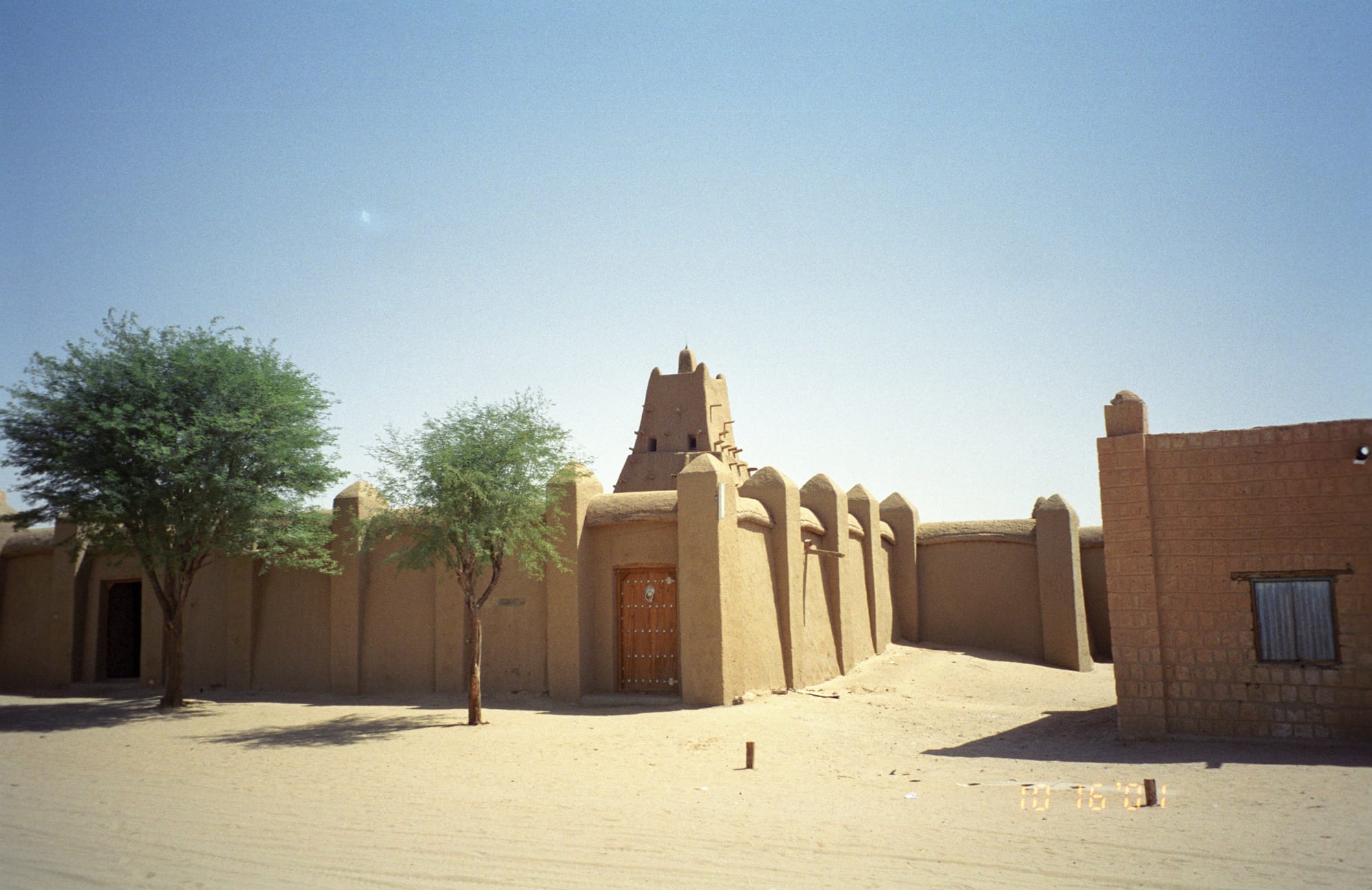 A mud-brick mosque with a tapering tower and buttressed walls on a sandy street, with two green trees in front
