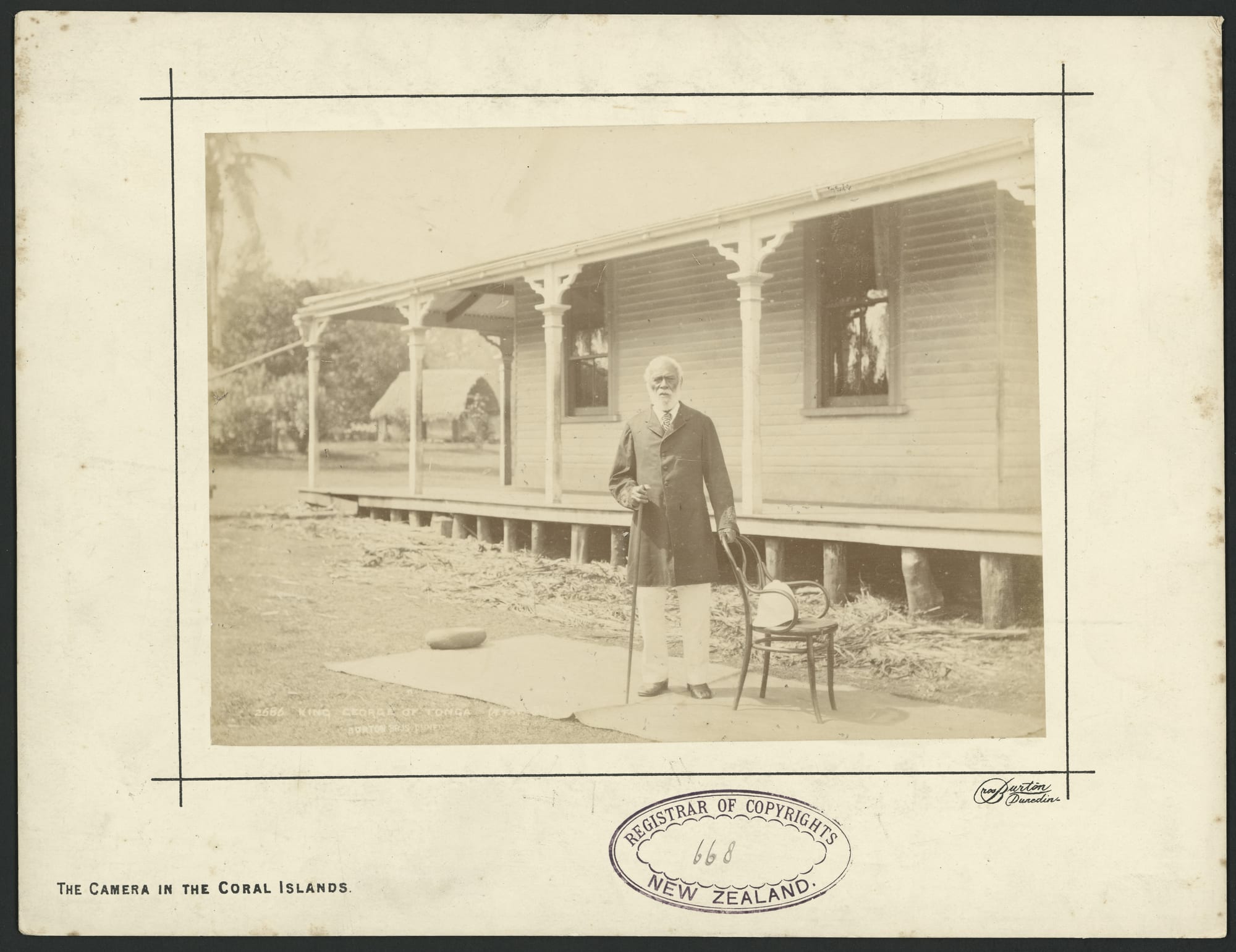 An old photograph of an elderly bearded man in a dark coat and white trousers standing with a cane beside a chair on a mat outside a veranda