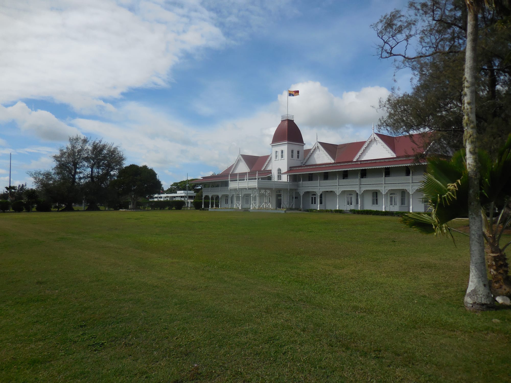 A white wooden palace with a red roof, verandas and a domed tower, flying a flag above a wide lawn