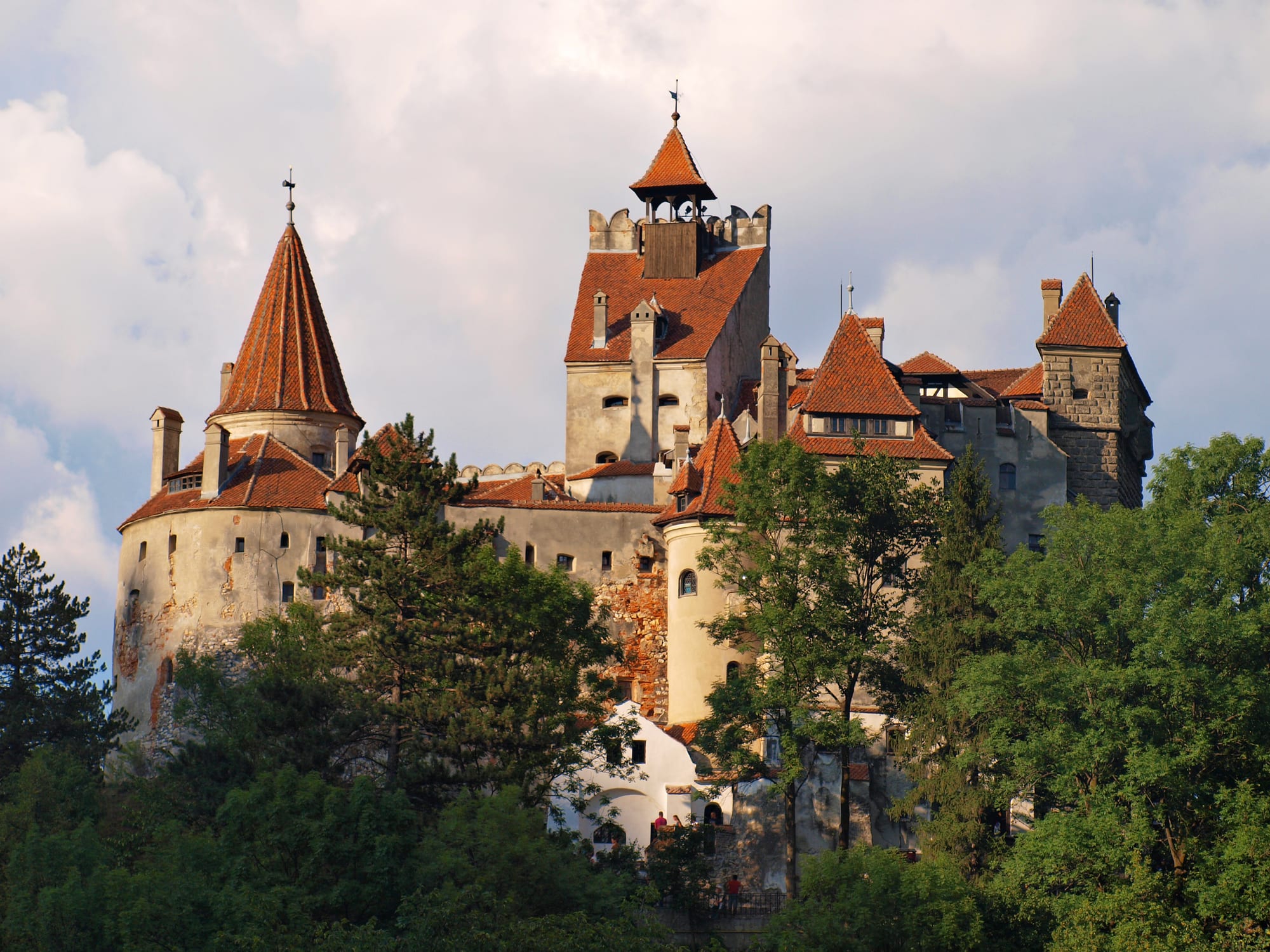 A pale stone castle with steep red-tiled roofs and towers rising above dark green trees under a cloudy sky