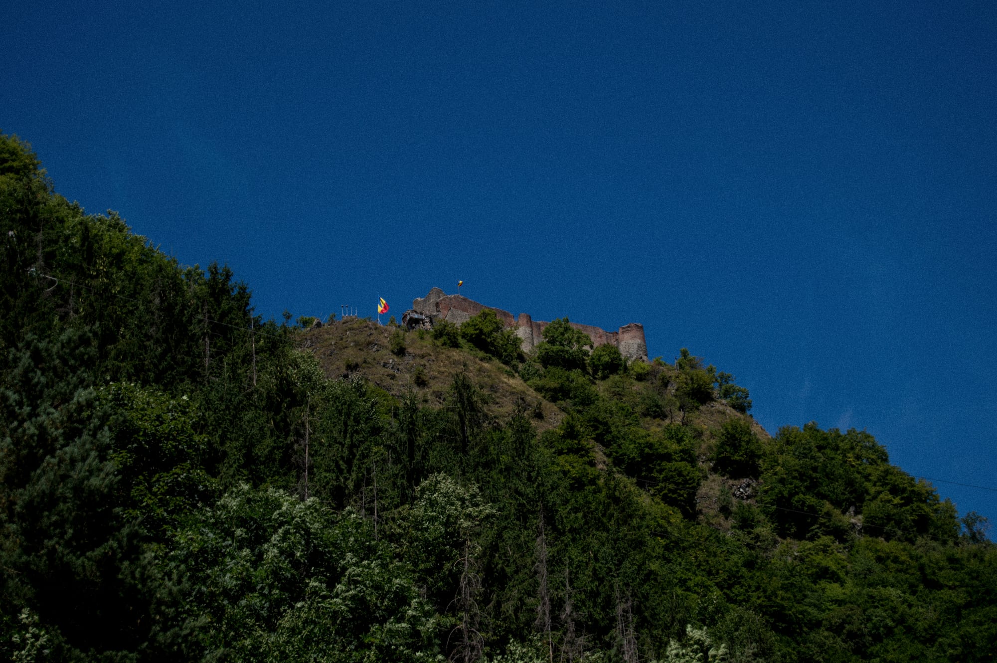 Ruined stone walls and towers on the summit of a steep forested mountain against a deep blue sky