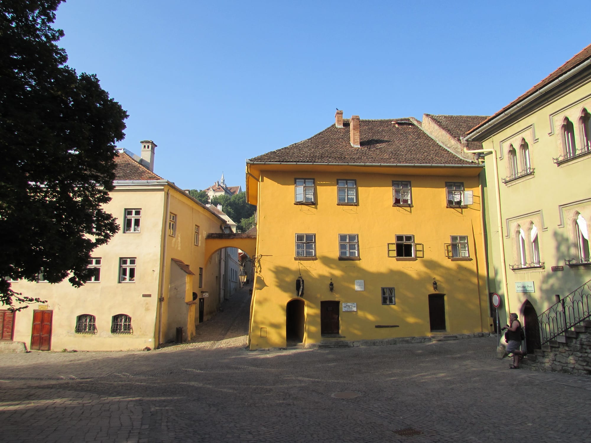 A mustard-yellow two-storey house with a steep tiled roof on a cobbled square, next to a pale arched gateway