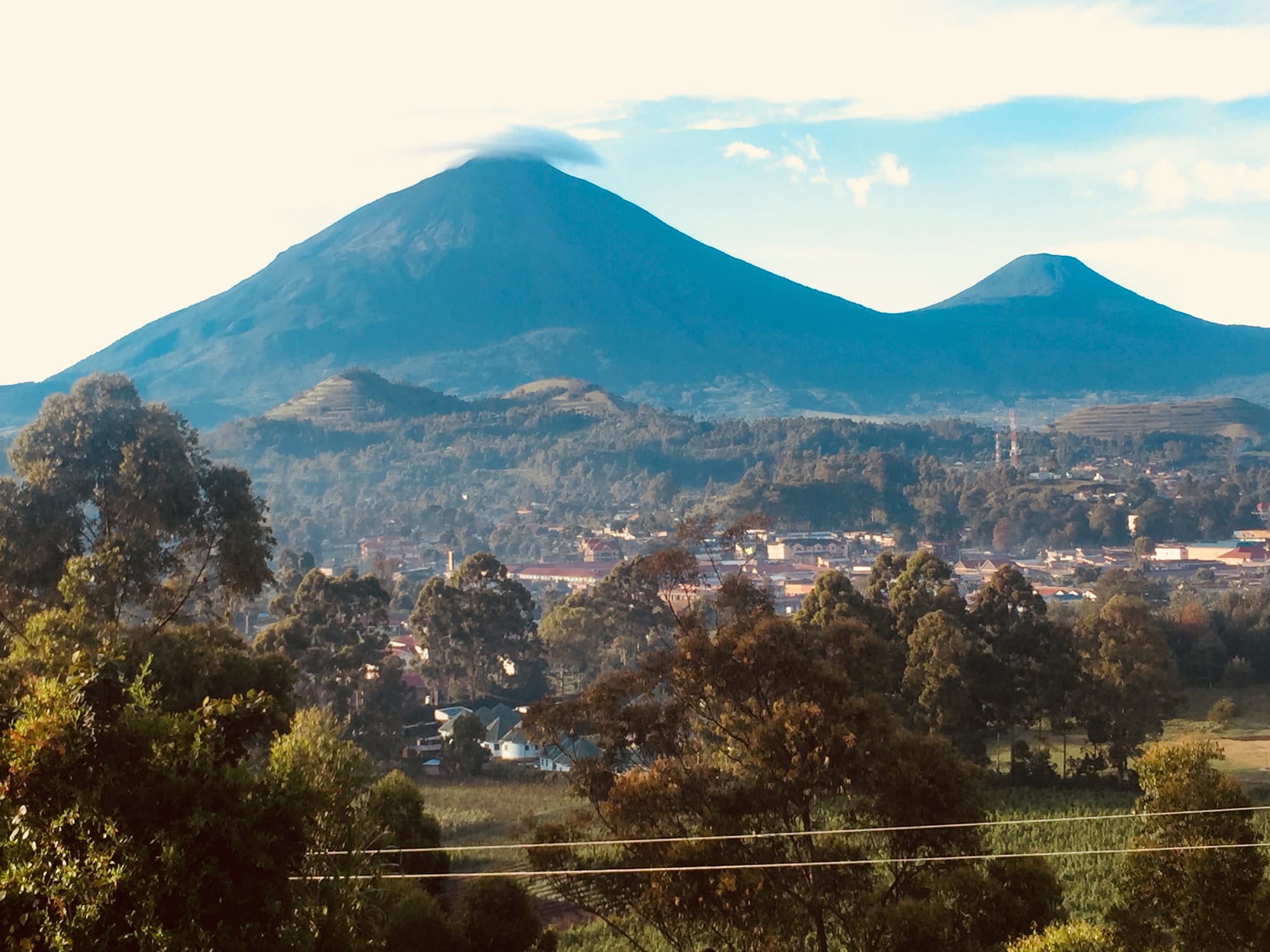 Two cone-shaped volcanoes, one taller with a cap of cloud, rising behind a small town and wooded farmland