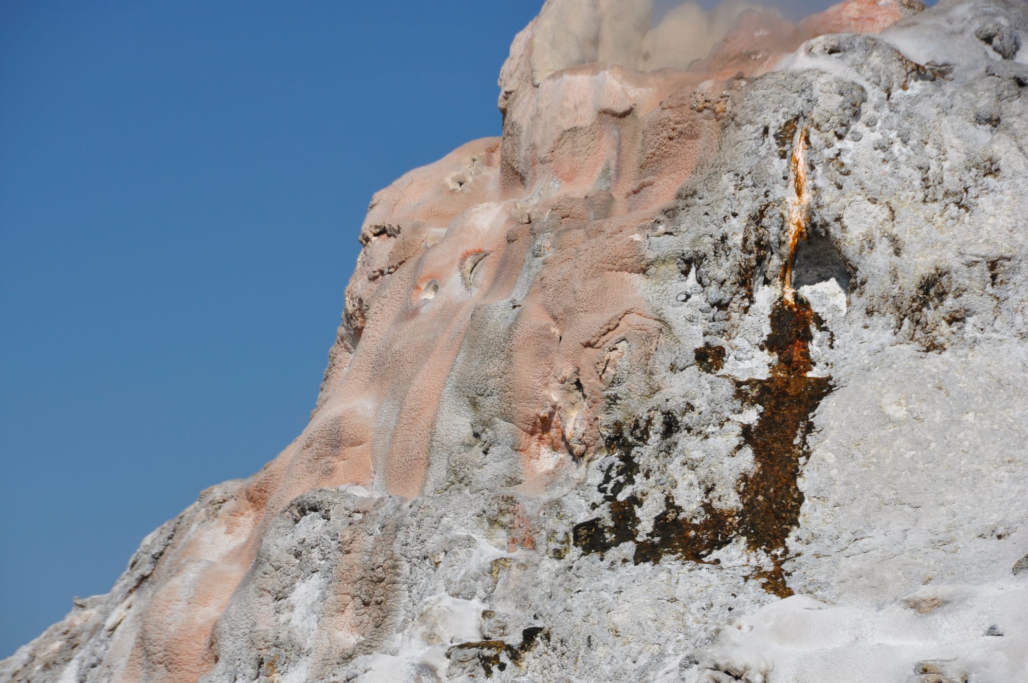 Close view of a rough grey, pink and rust-streaked mineral mound against a deep blue sky, with steam at its top
