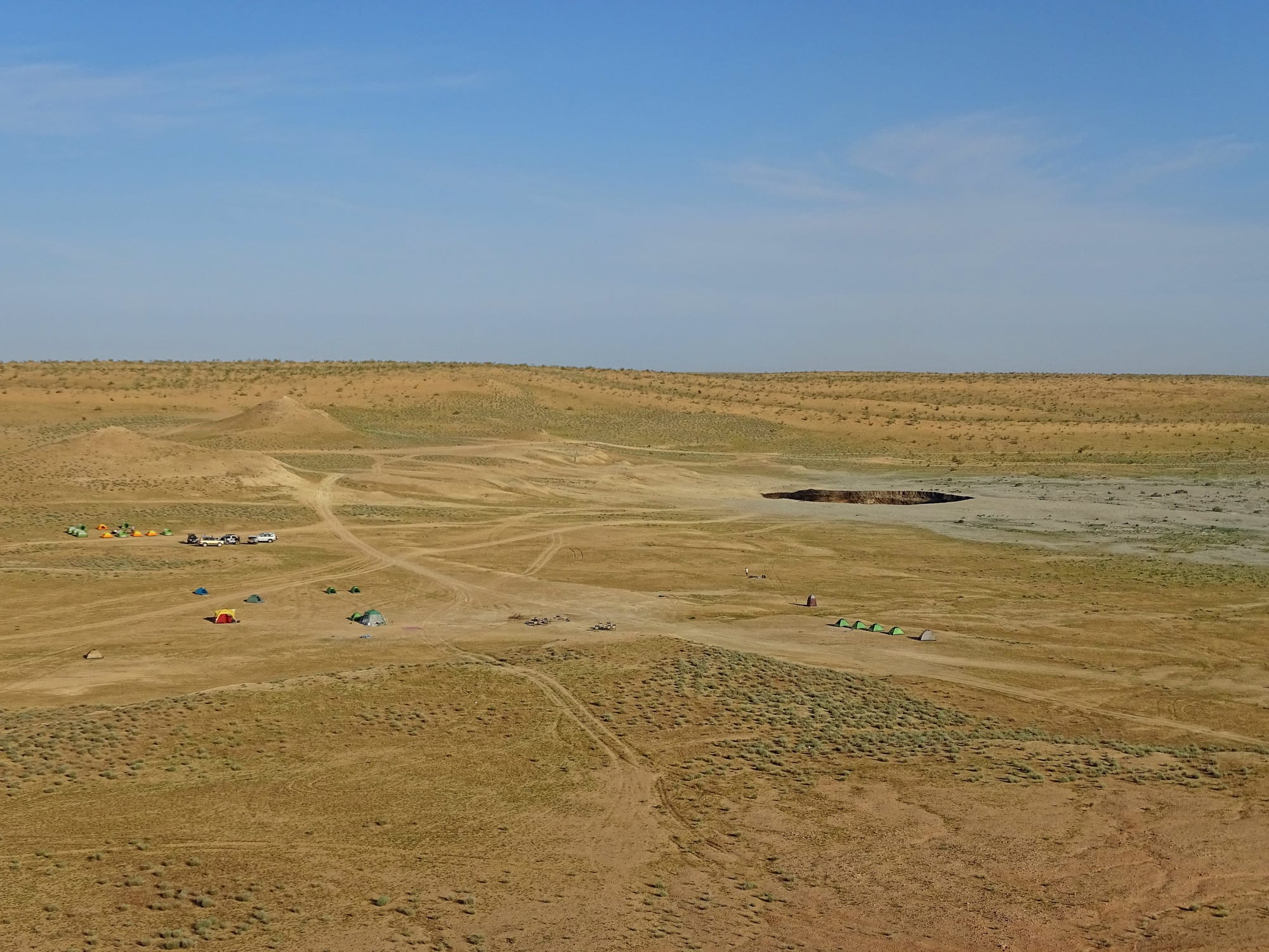 A wide view of flat sandy desert with scattered tents and parked cars, and a dark oval pit in the distance
