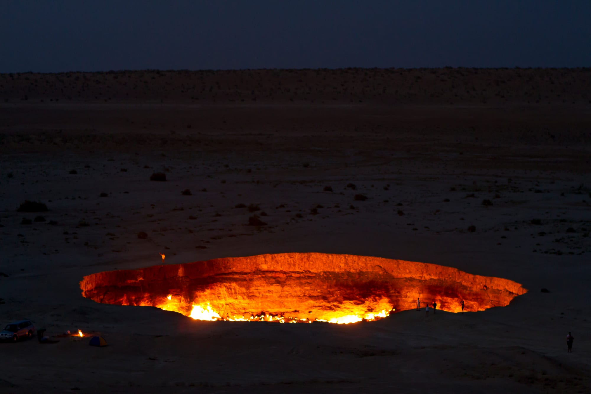 A wide oval pit glowing orange with flames along its floor in a dark, flat desert at dusk, with tiny figures standing at the rim